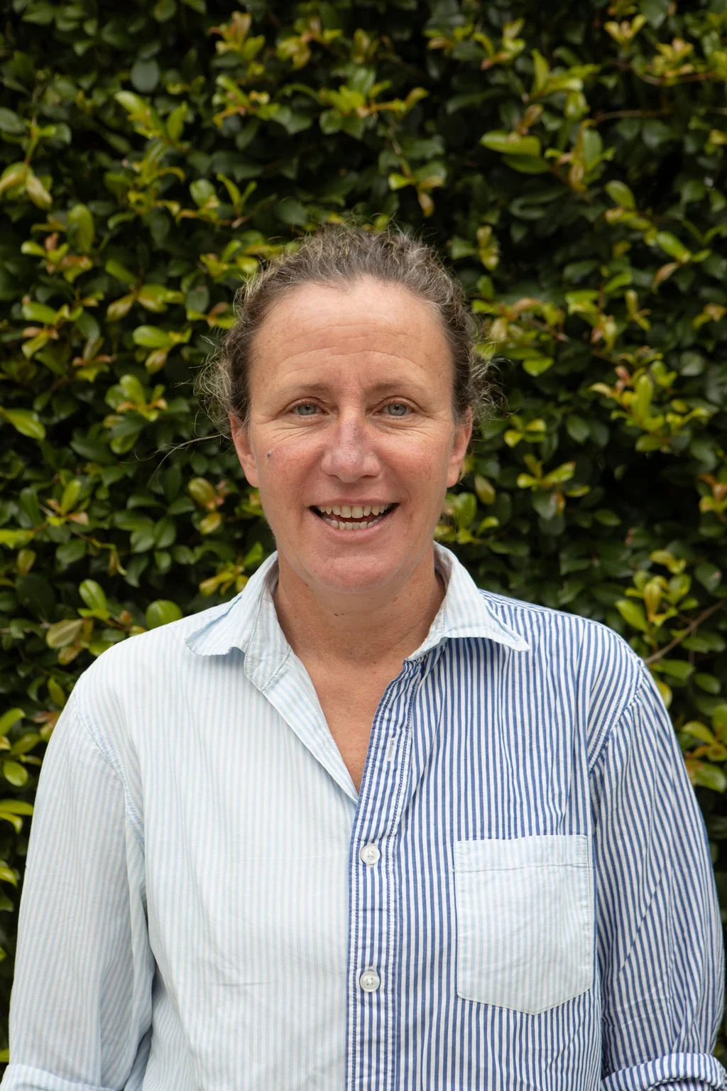 A smiling woman with light-colored hair pulled back, wearing a light blue and white vertically striped shirt, standing outdoors in front of a dense green hedge.