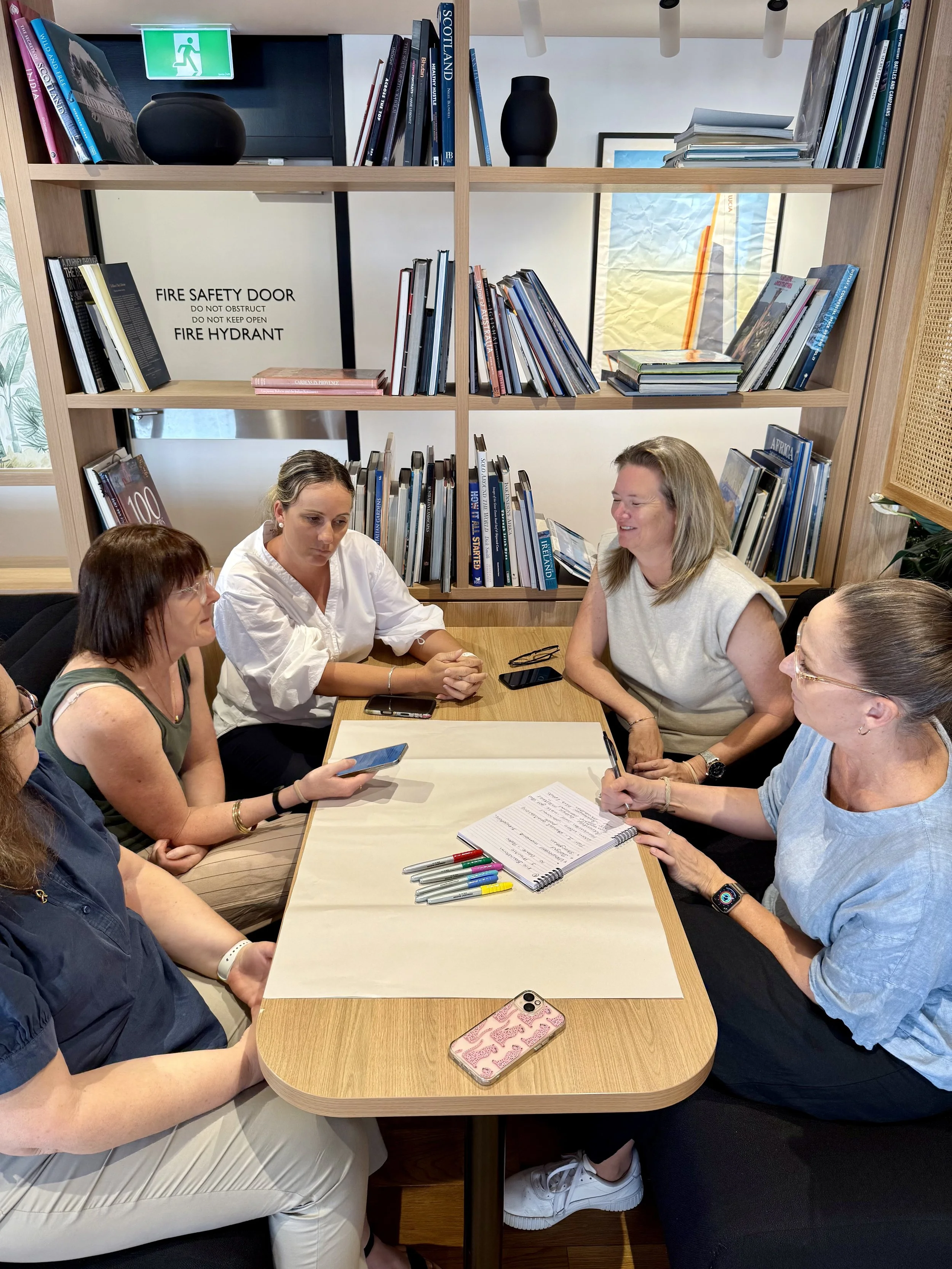Five women are sitting around a rectangular table engaged in a discussion. The table has a notepad with handwritten notes, colorful pens, and a smartphone. A bookshelf with many books and a safety sign are visible in the background.