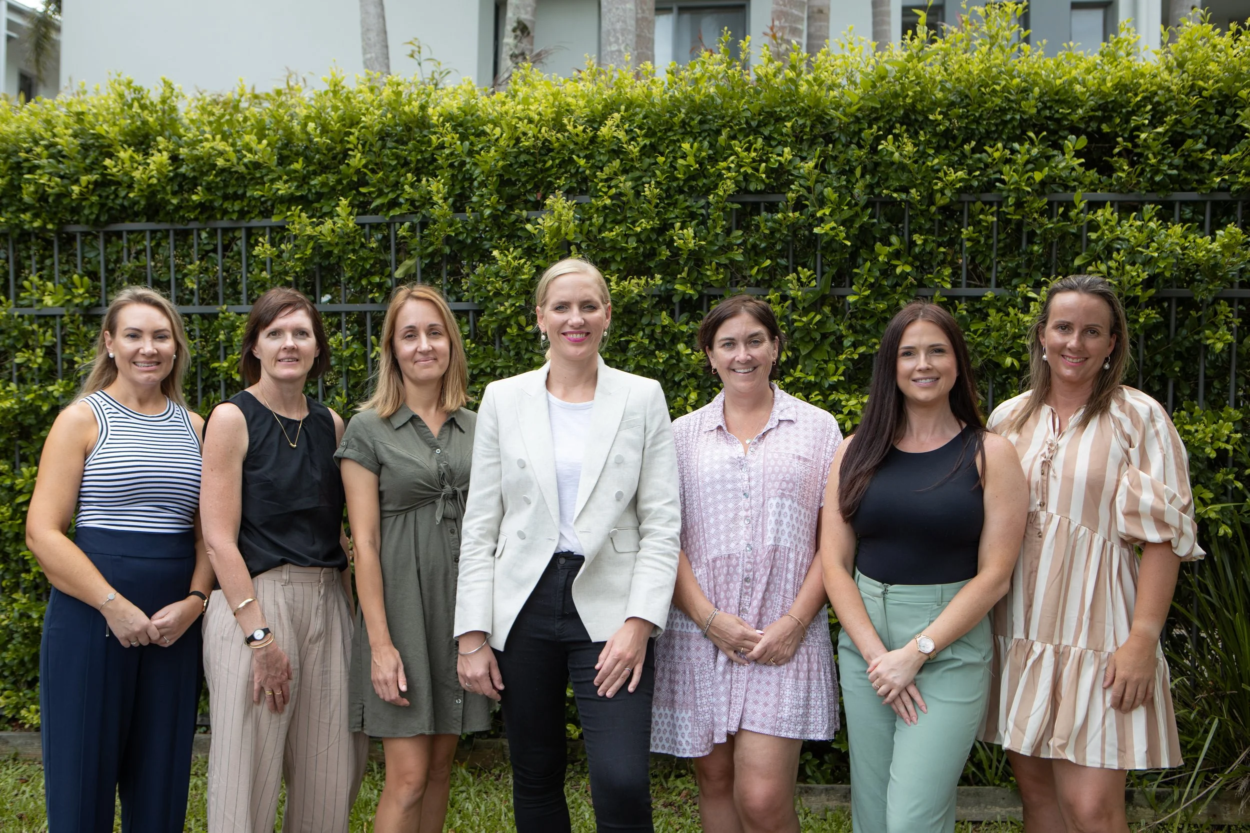 Group of seven women standing outdoors in front of a green hedge, smiling at the camera.