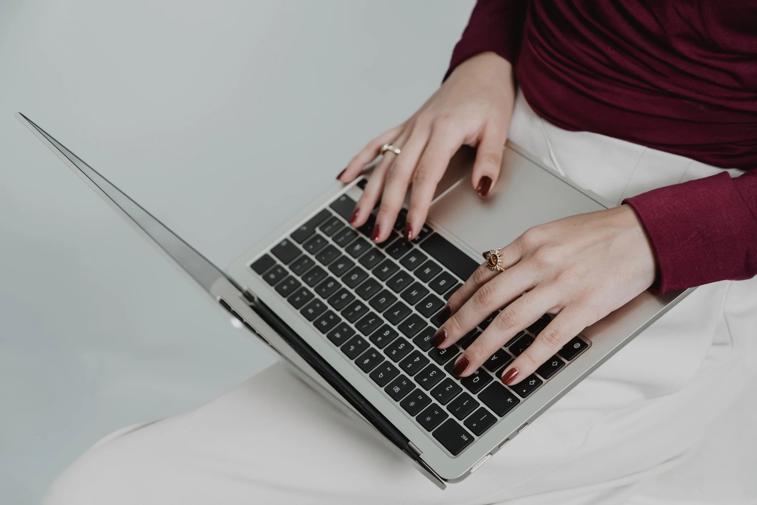 Manos de mujer con uñas color burdeos y anillos, tecleando en un portátil plateado, con ropas color vino y blancas