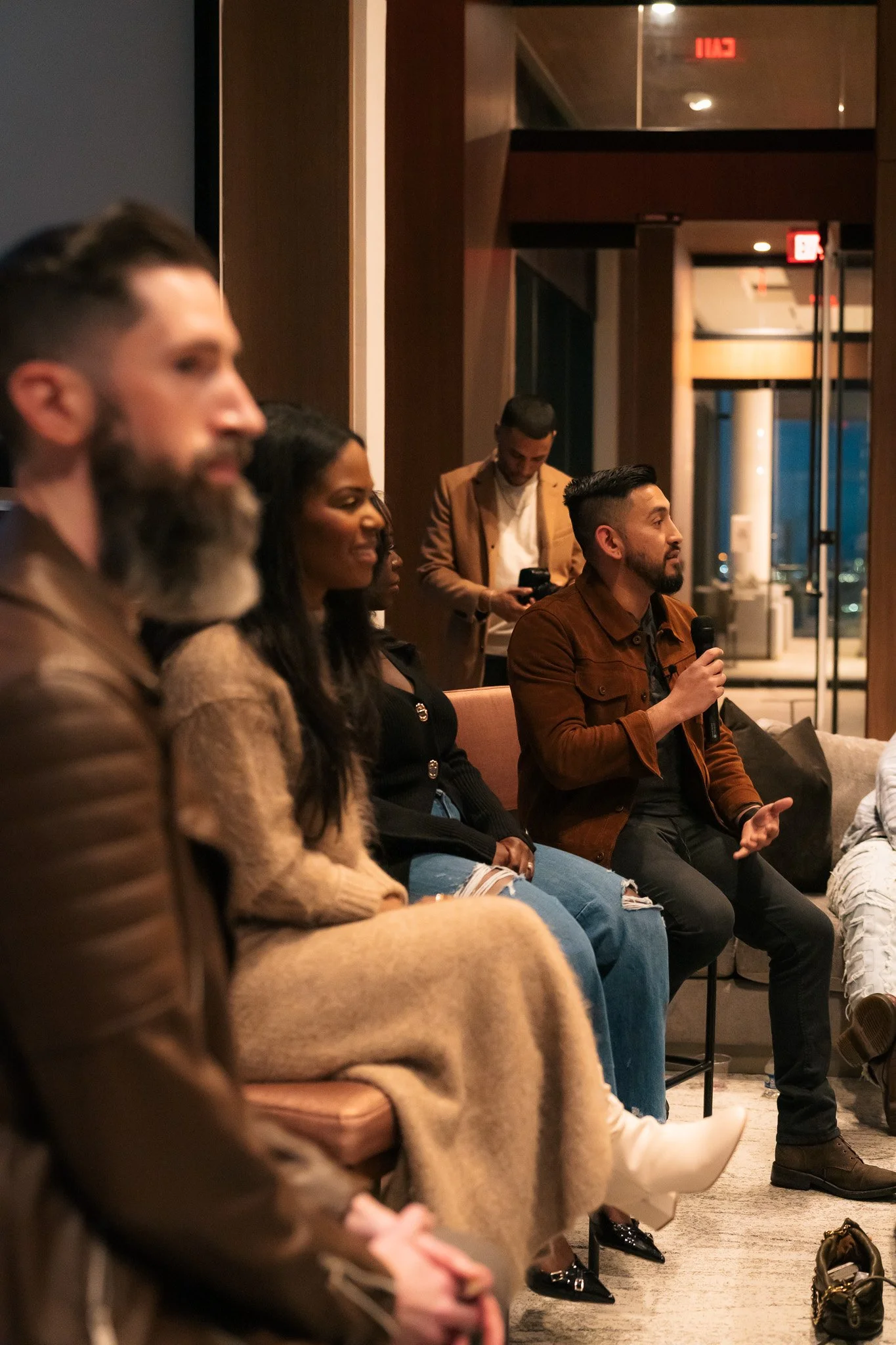 Group of diverse people sitting on a couch in a modern indoor setting, engaging in a discussion or presentation, with one person holding a microphone.