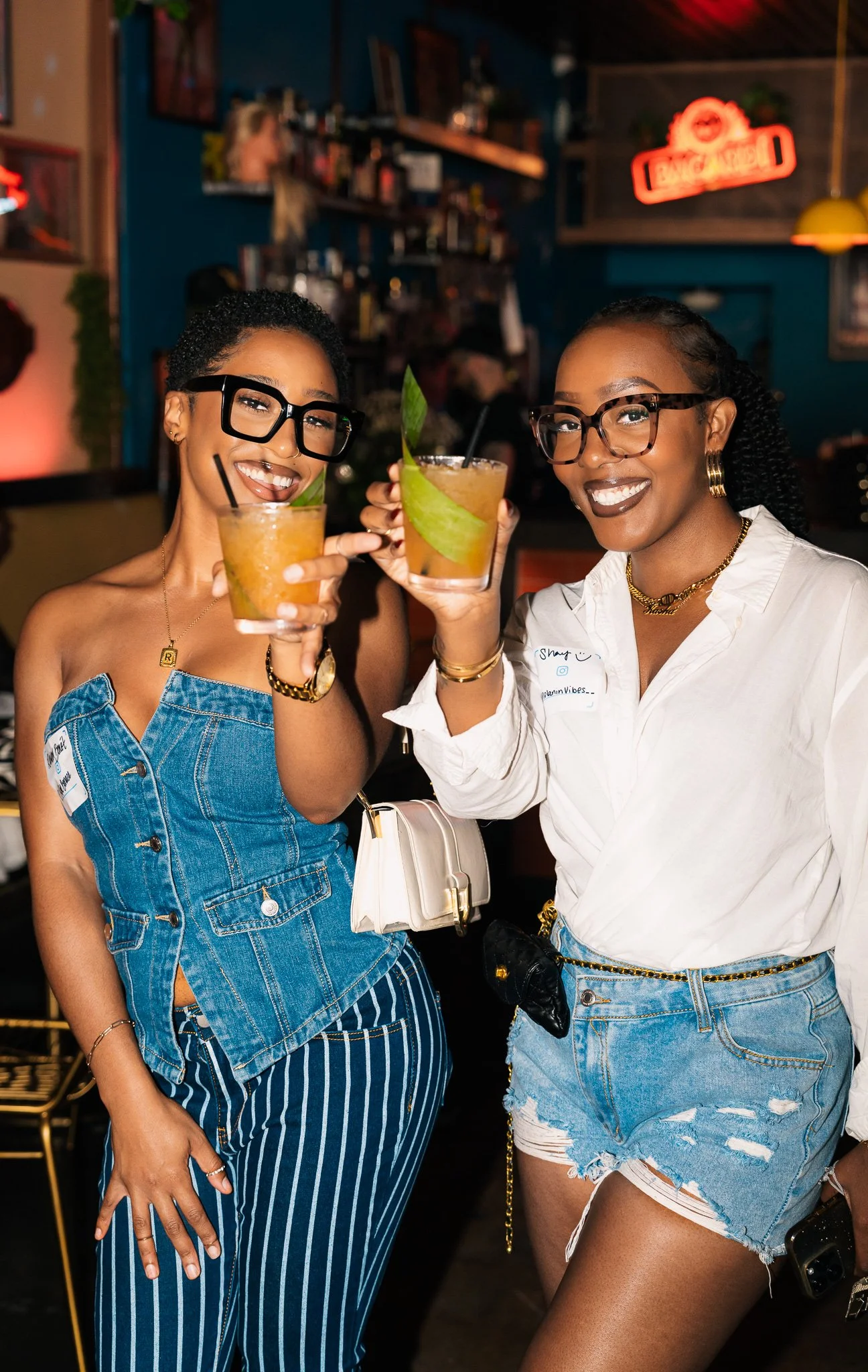 Two women smiling and holding drinks in a bar or restaurant with a neon sign in the background.