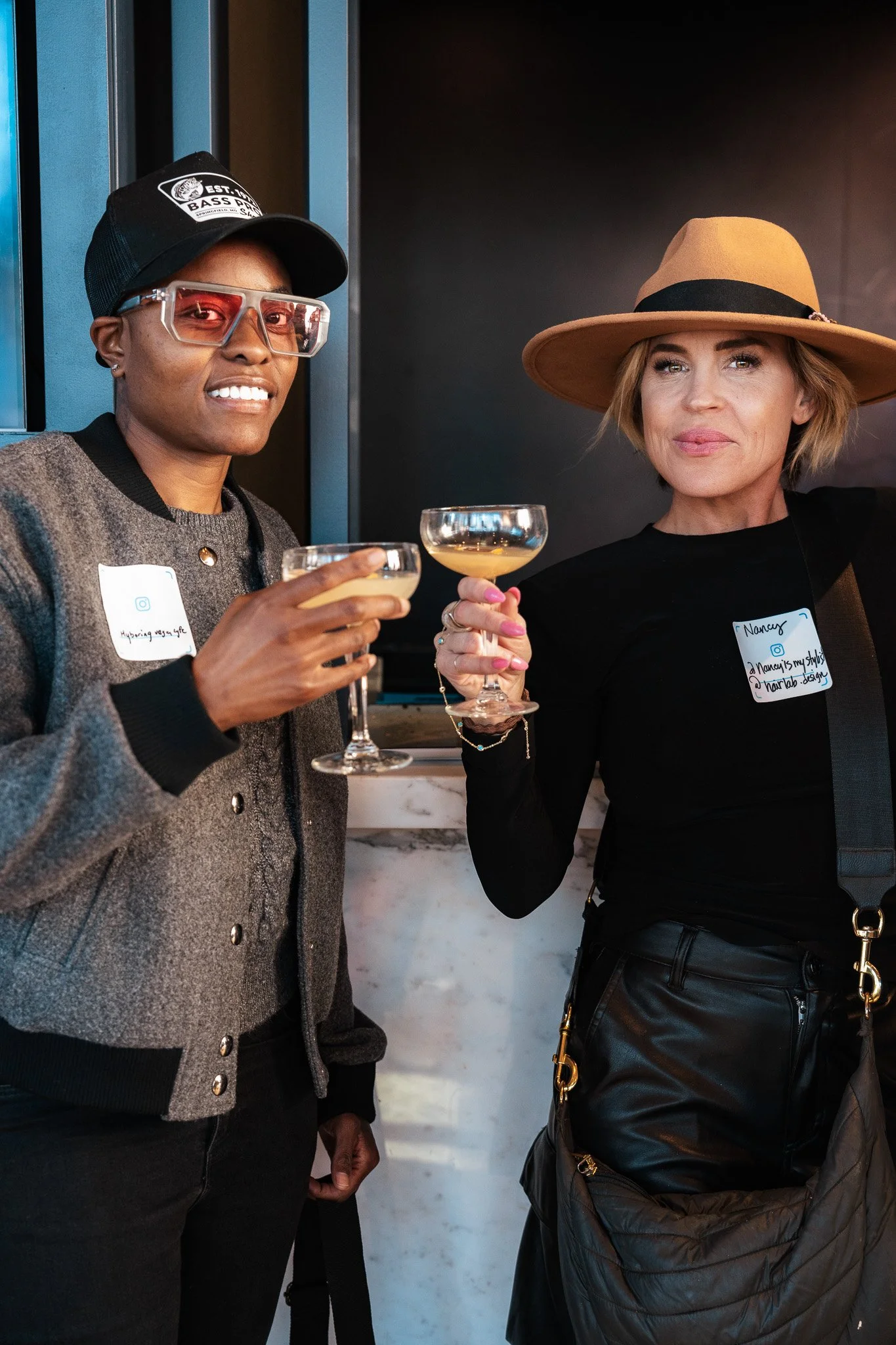 Person in a shiny pink sequined jacket, matching pink pants, white furry hat, and white chunky sneakers, posing indoors with a confident smile, others in the background taking photos.