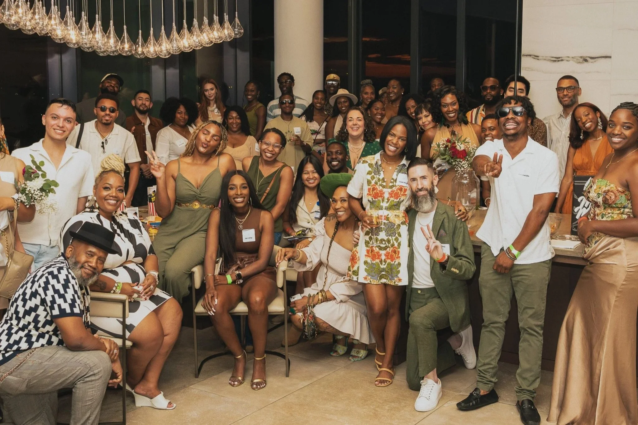 A large group of diverse people gathered indoors at a social event, smiling and posing for the camera, with some making peace signs, and a chandelier hanging from the ceiling.
