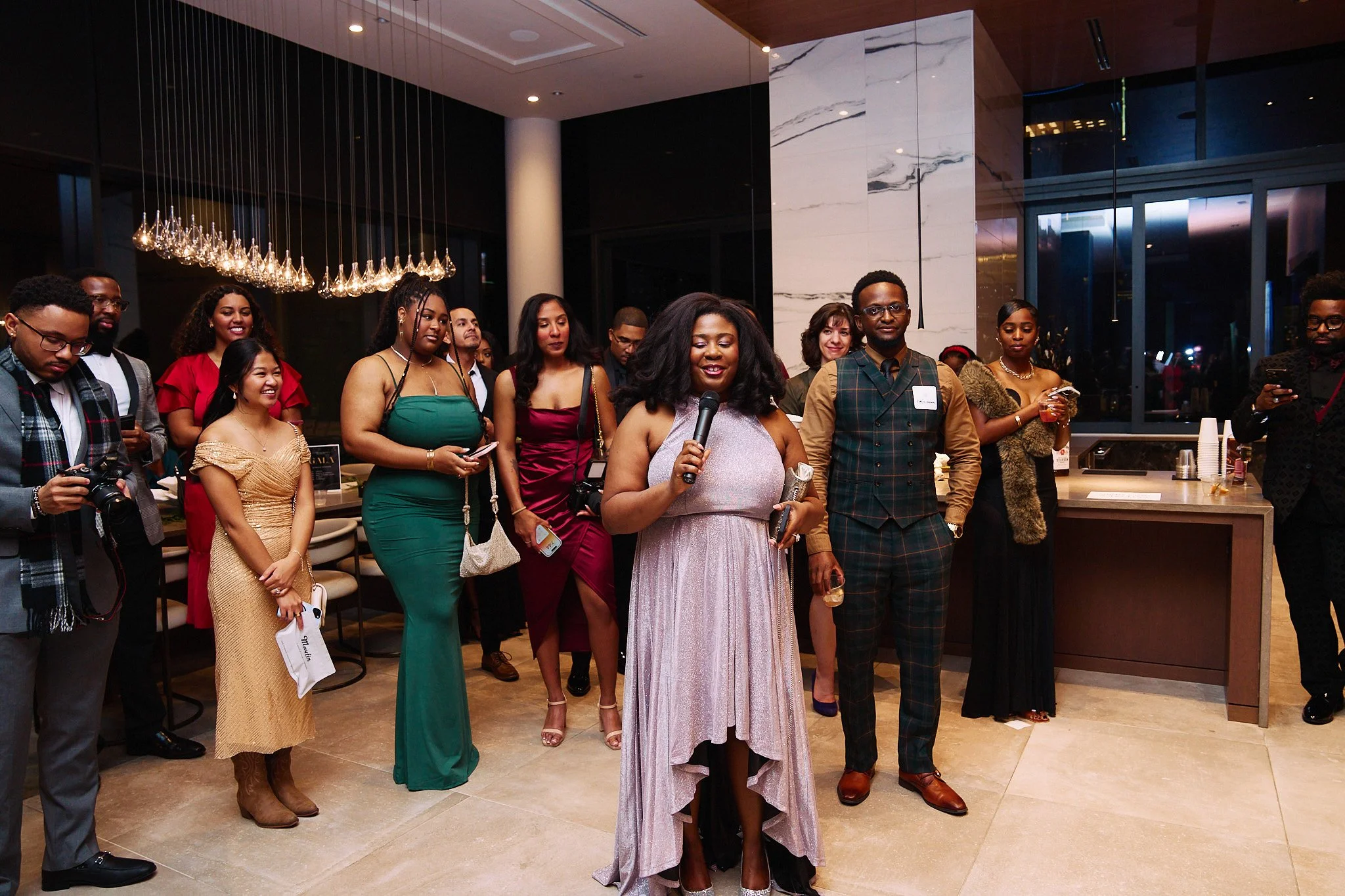 A group of people dressed in formal attire gathered at an indoor event, with a woman in a lavender dress holding a microphone speaking to the crowd.