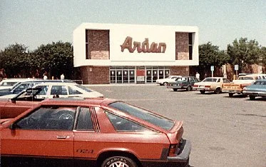 A shopping plaza with a sign displaying the word 'Adam' and several parked cars in front of the store.