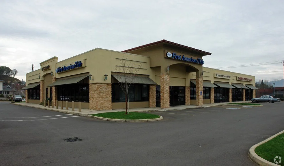A strip mall with a beige building featuring multiple storefronts, including one for First American Title, with parking lot and small landscaped areas in front and on the side, under an overcast sky.