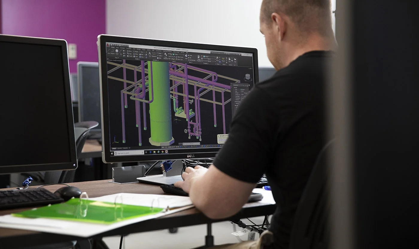 A person sitting at a desk working on a CAD design of an industrial piping system on a computer monitor.