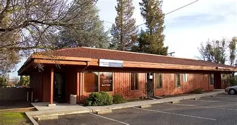 A single-story brick building with a red-tiled roof and four windows, located in a parking lot with a few parked cars and surrounded by trees.