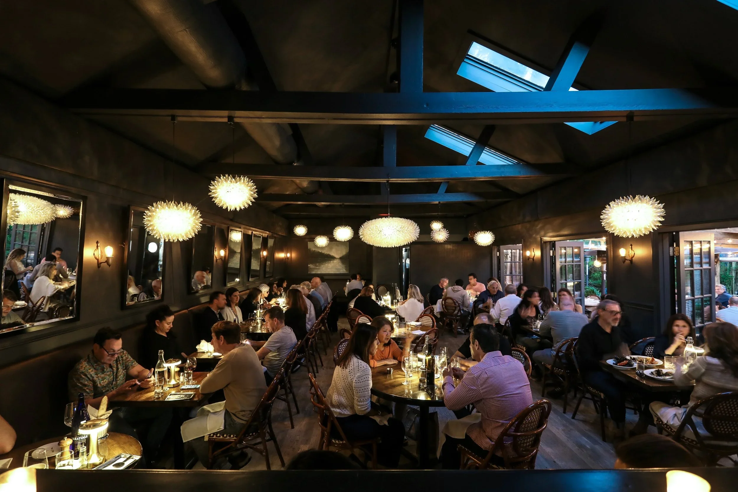 Interior of a busy restaurant with people dining at tables, modern chandeliers, and windows open to outdoor seating.