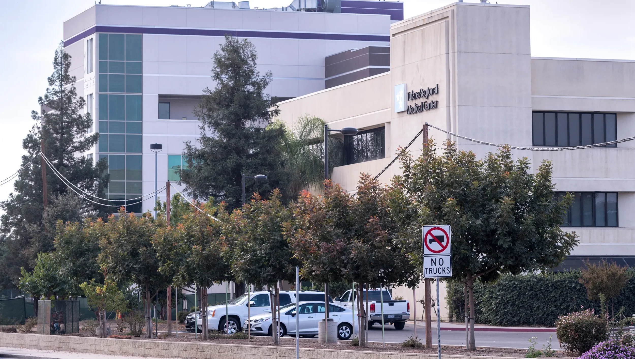 Exterior view of Tulare Regional Medical Center, a modern multi-story hospital building with large glass windows, surrounded by parked cars, trees, and a 'No Trucks' traffic sign.