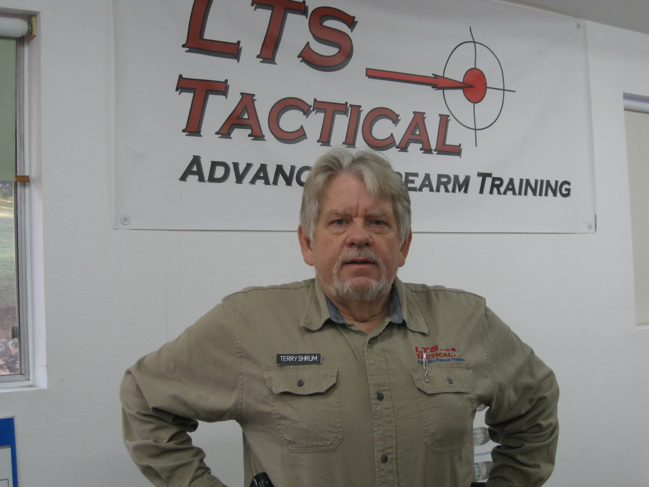 Man wearing a beige shirt with a name tag that says 'Terry Shrum' standing indoors in front of a banner that reads 'LTS Tactical' and 'Advanced Firearm Training' with a target symbol.