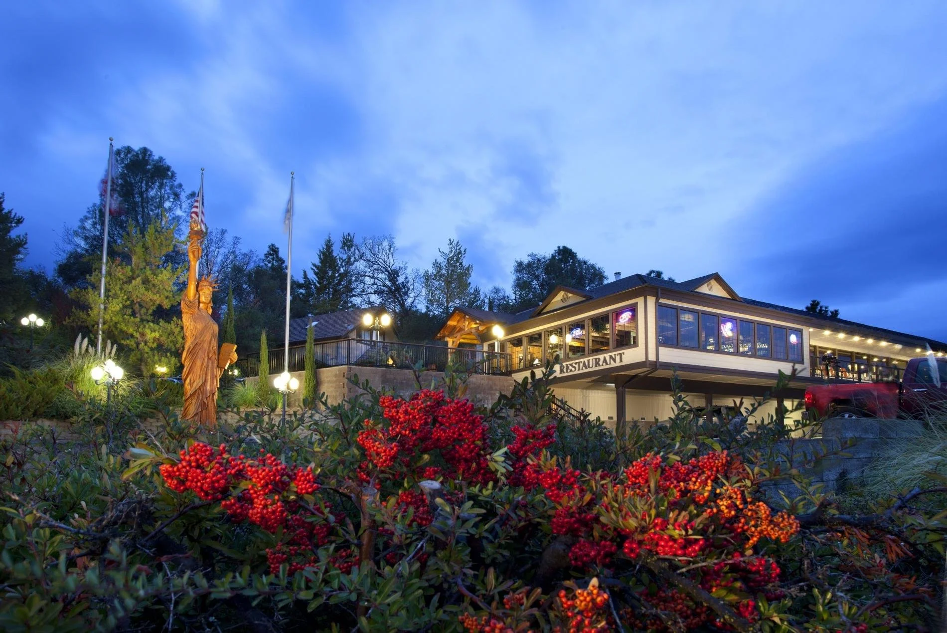 A restaurant building with large windows illuminated from inside, situated on a slight hill. In the foreground, there are red berries and green foliage. To the left, a carved wooden statue of the Statue of Liberty and three flagpoles with flags stand near illuminated garden lights. The sky above is transitioning from dusk to night.