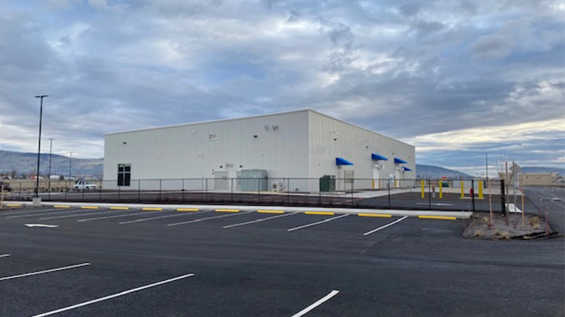 Empty parking lot with white industrial building in the background under cloudy sky.