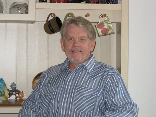 A man with gray hair and a beard, wearing a striped blue and white shirt, standing in a kitchen with hanging mugs and a white cabinet.