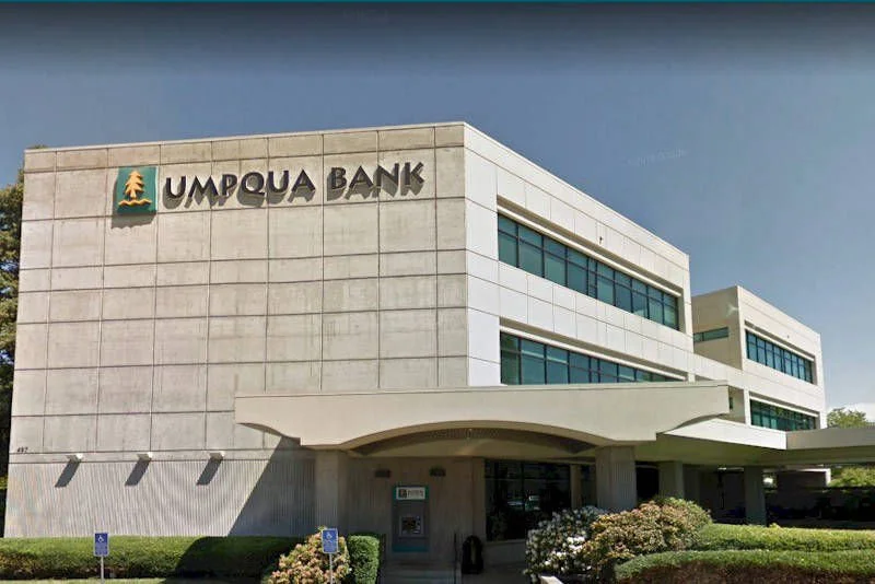 Umpqua Bank building with beige concrete facade and large windows, green bushes and handicap parking signs in front, under a clear blue sky.
