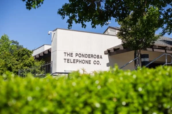 White building with black text that reads 'The Ponderosa Telephone Co.' partly obscured by green bushes and trees.