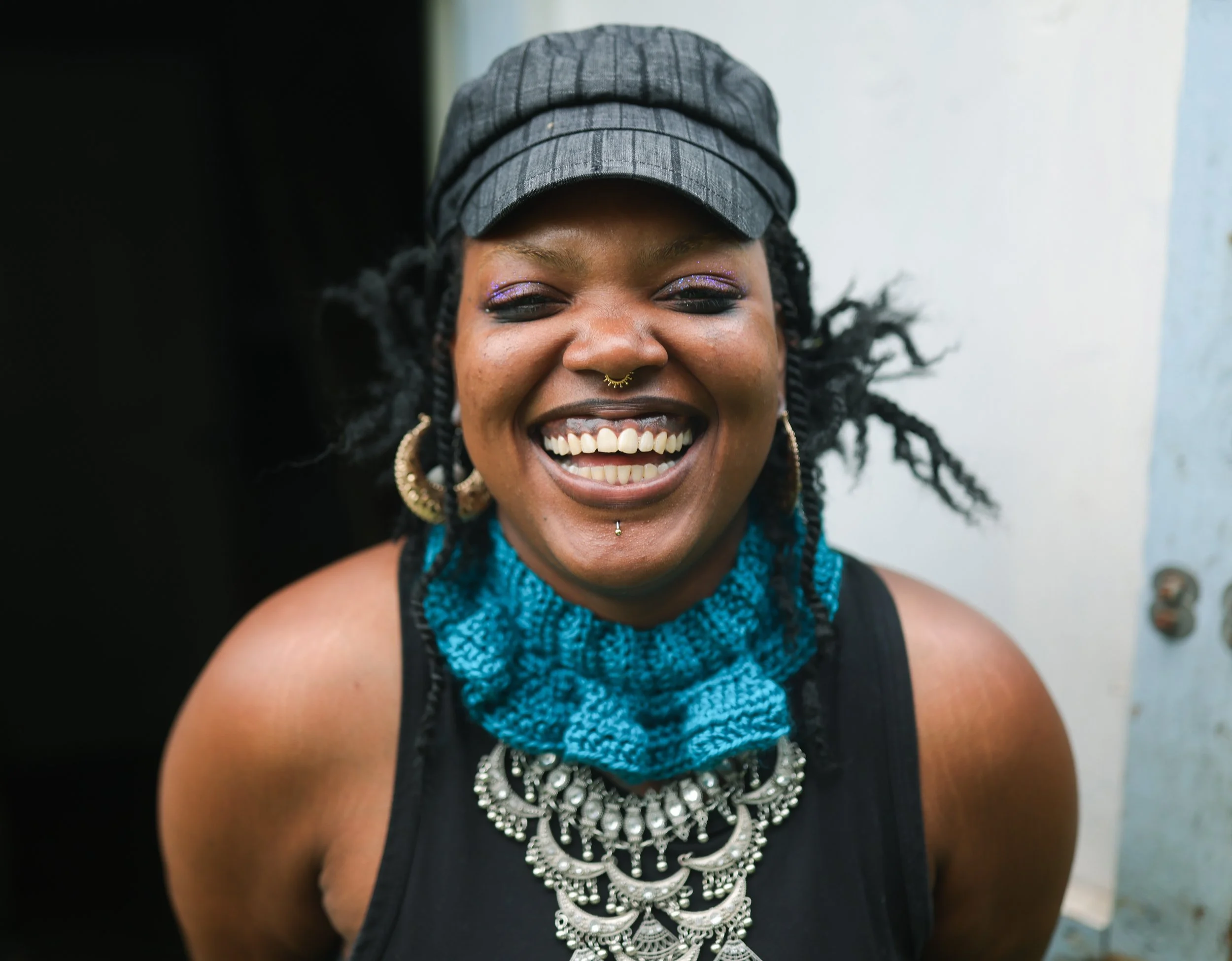 A smiling woman with dark skin, curly hair, and colorful makeup, wearing a black tank top, a gray cap, a blue textured scarf, large earrings, and a statement necklace.