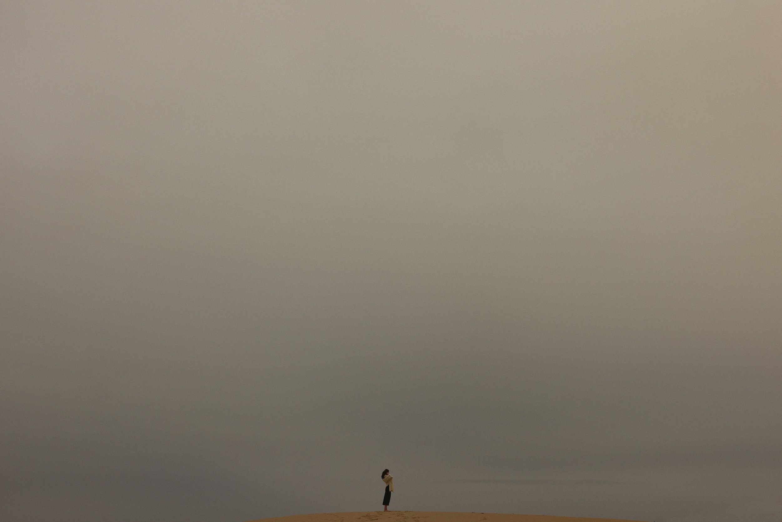 A person standing alone on a sandy hill under a large, overcast sky.