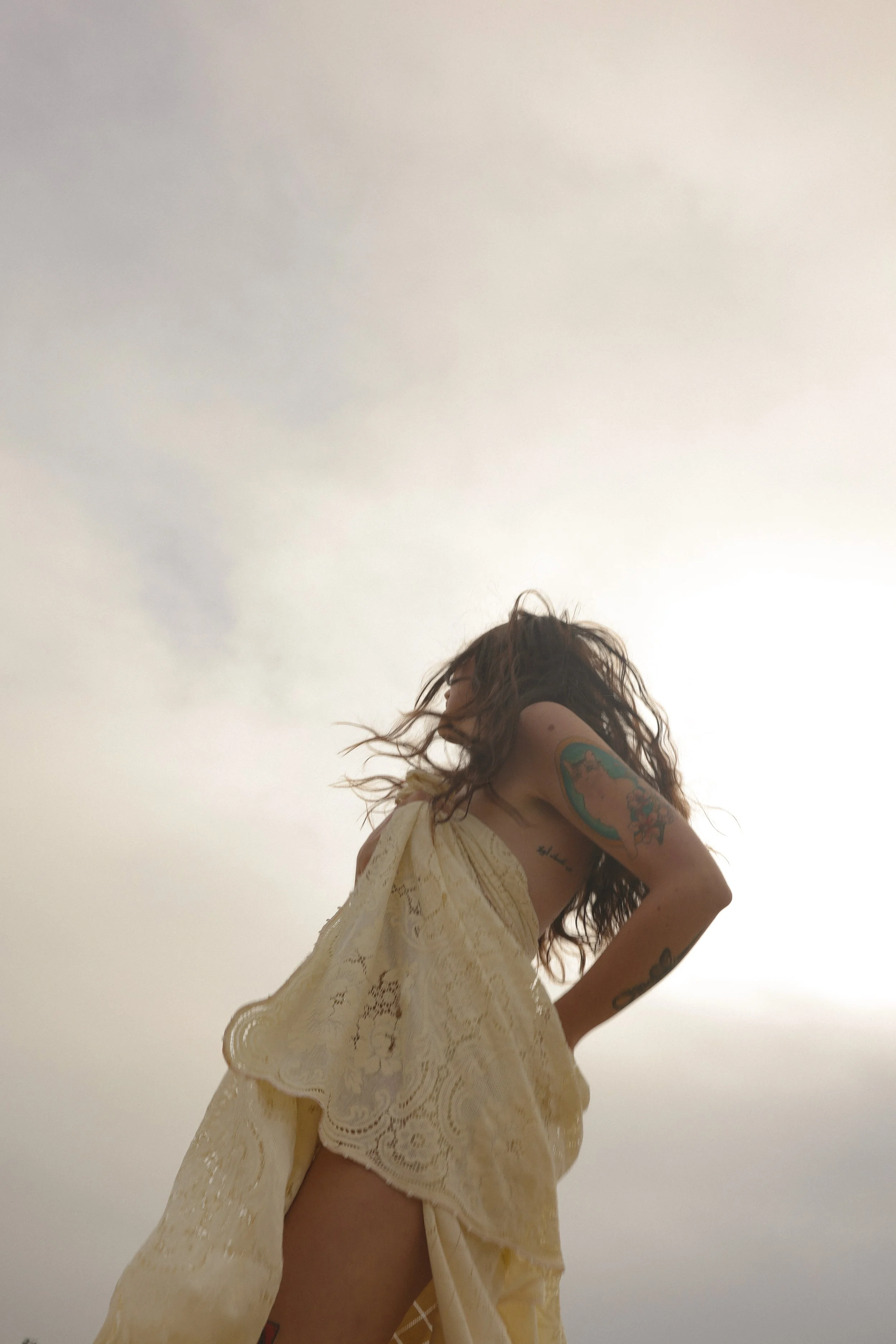 A woman with long curly hair standing outdoors against a cloudy sky, wearing a beige lace dress, with tattoos on her arm.