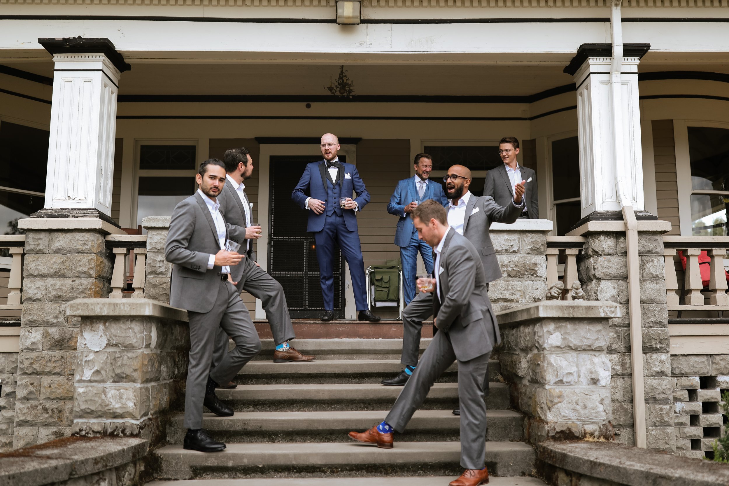 Group of men in suits socializing on the steps of a porch outside a house, holding drinks and engaged in conversation.