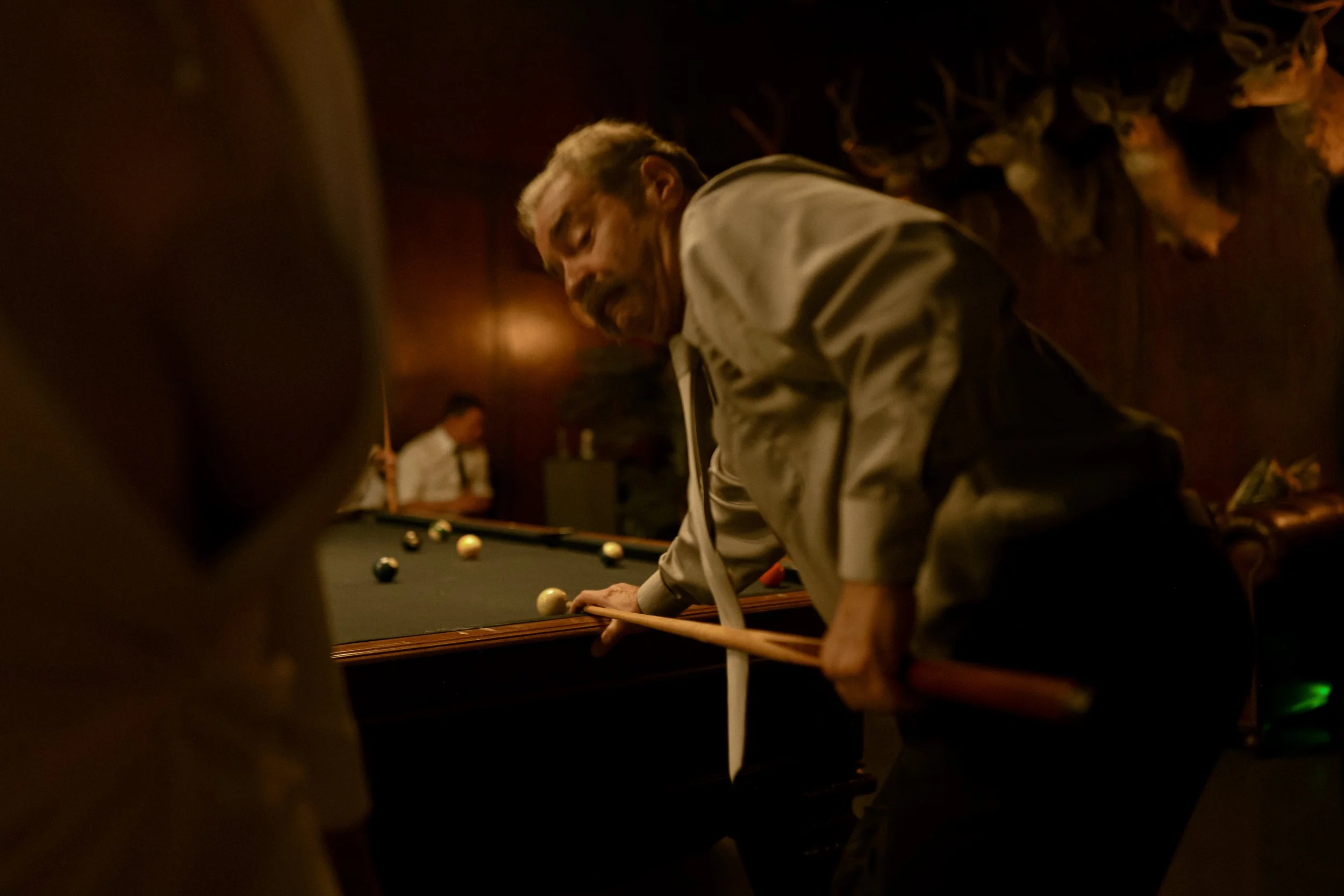 A man with gray hair and a mustache, wearing a light-colored shirt, is playing billiards in a dimly lit room. He is leaning over the pool table, aiming to take a shot with a cue stick. Another person in a white shirt is visible in the background, hol