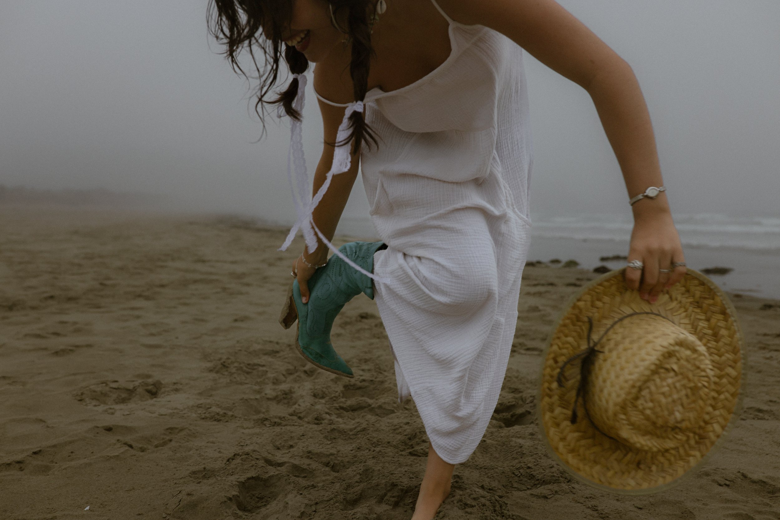 A woman with braids smiles while on a foggy beach, holding a straw hat in one hand and a boot in the other, dressed in a white dress.