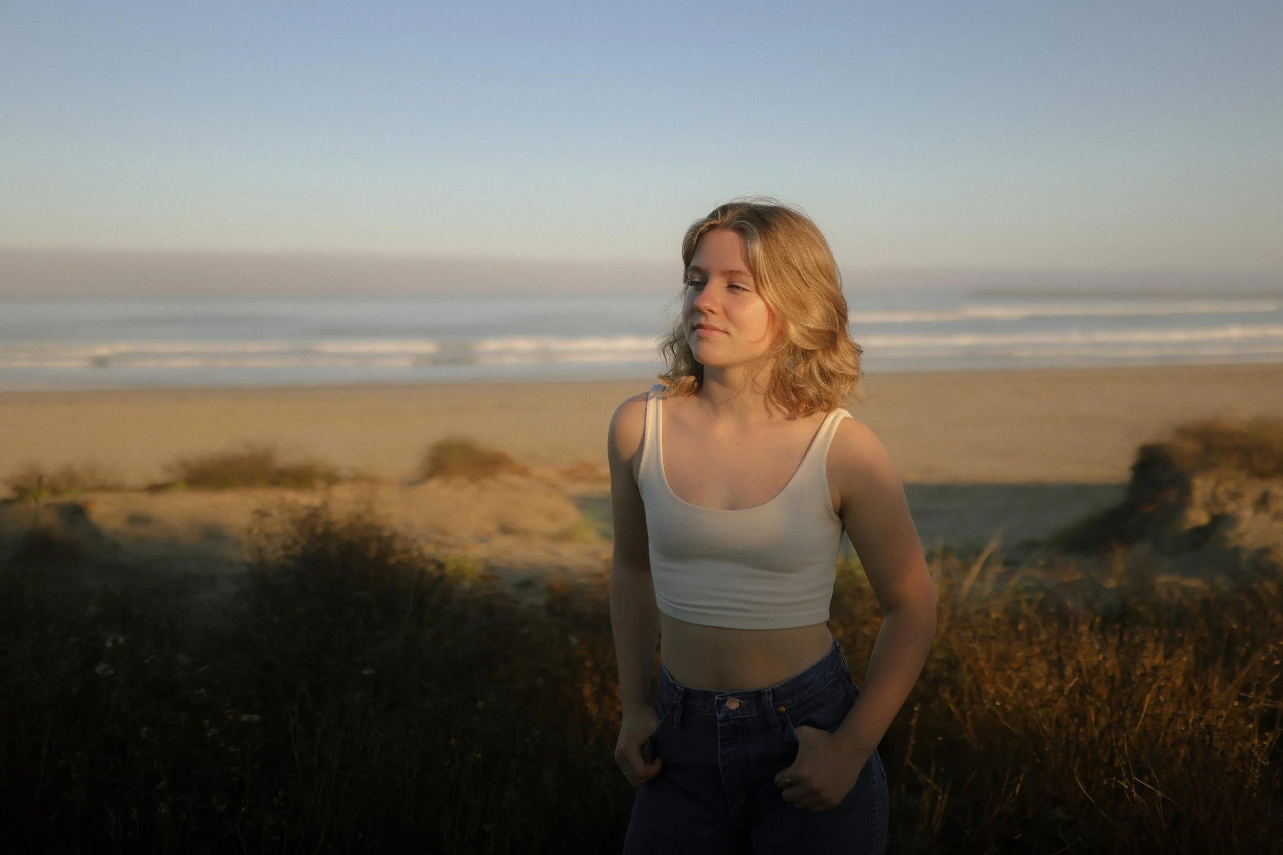 A young woman with shoulder-length blonde hair standing on a beach during sunset, wearing a white crop top and dark jeans, with the ocean and sky in the background.