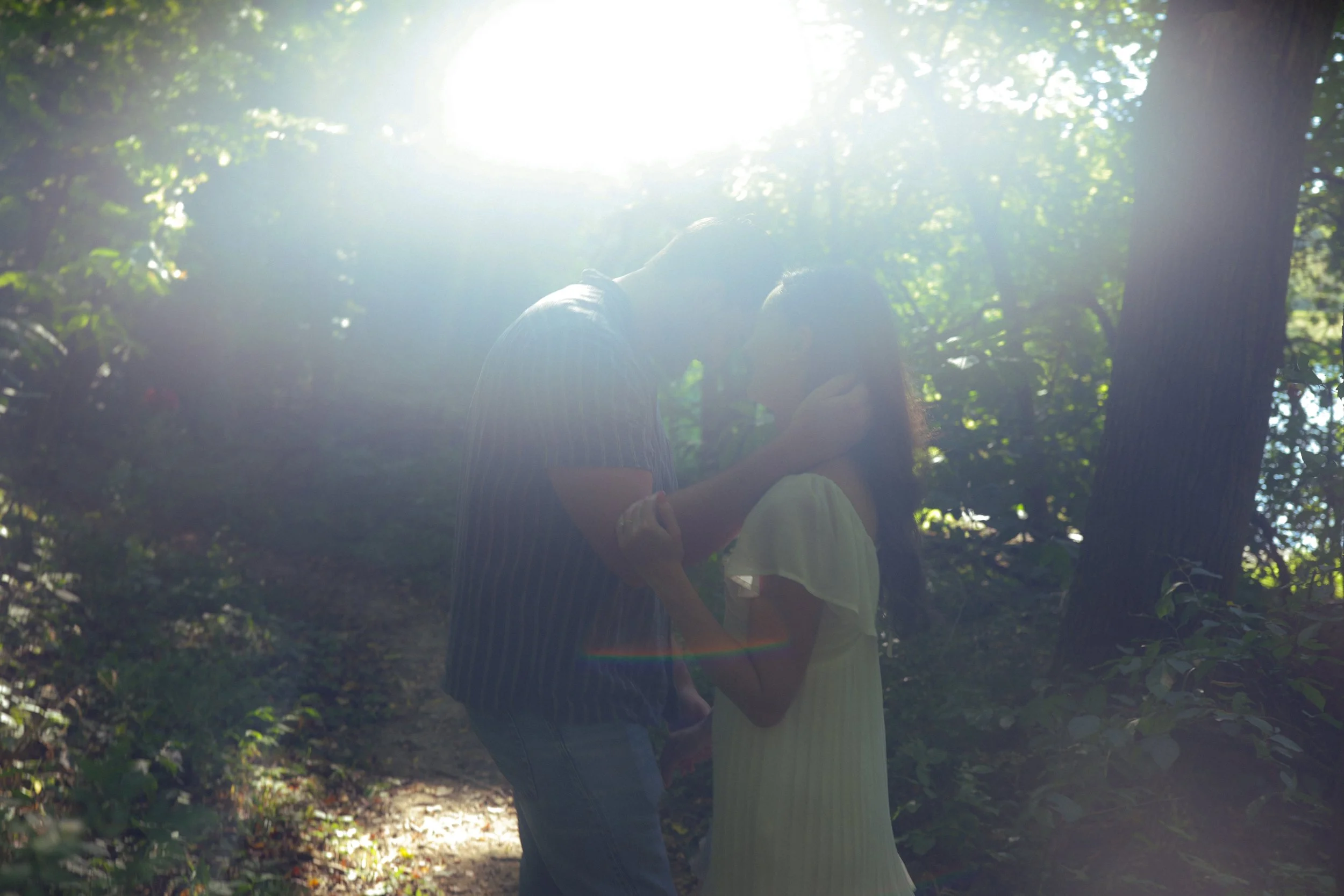 A couple stands close together in a wooded area, holding hands and touching foreheads, with sunlight filtering through the trees behind them.
