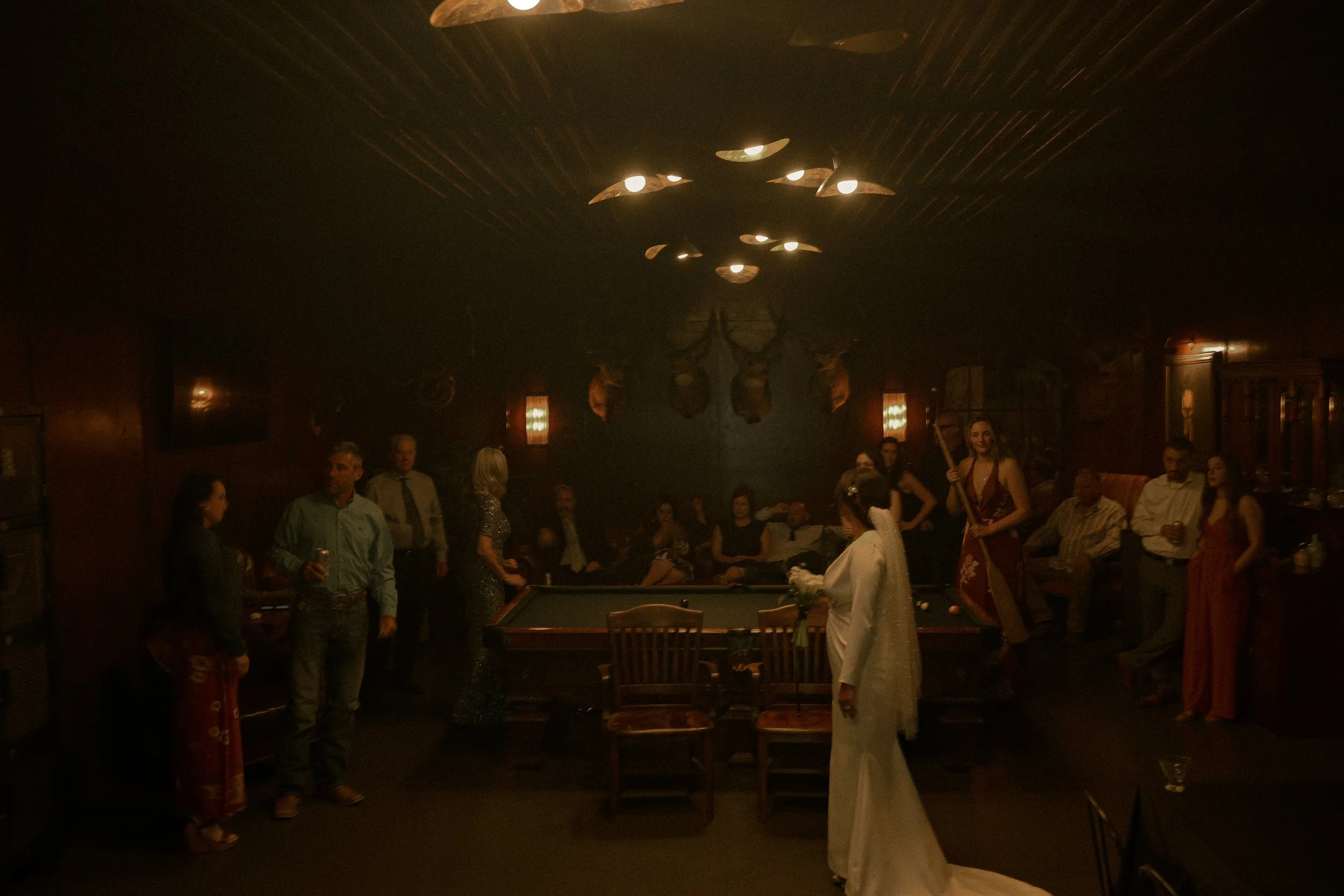 A group of people socializing in a dimly lit bar or lounge with a woman in white traditional attire in the foreground, and decorative mounted animal heads on the back wall.