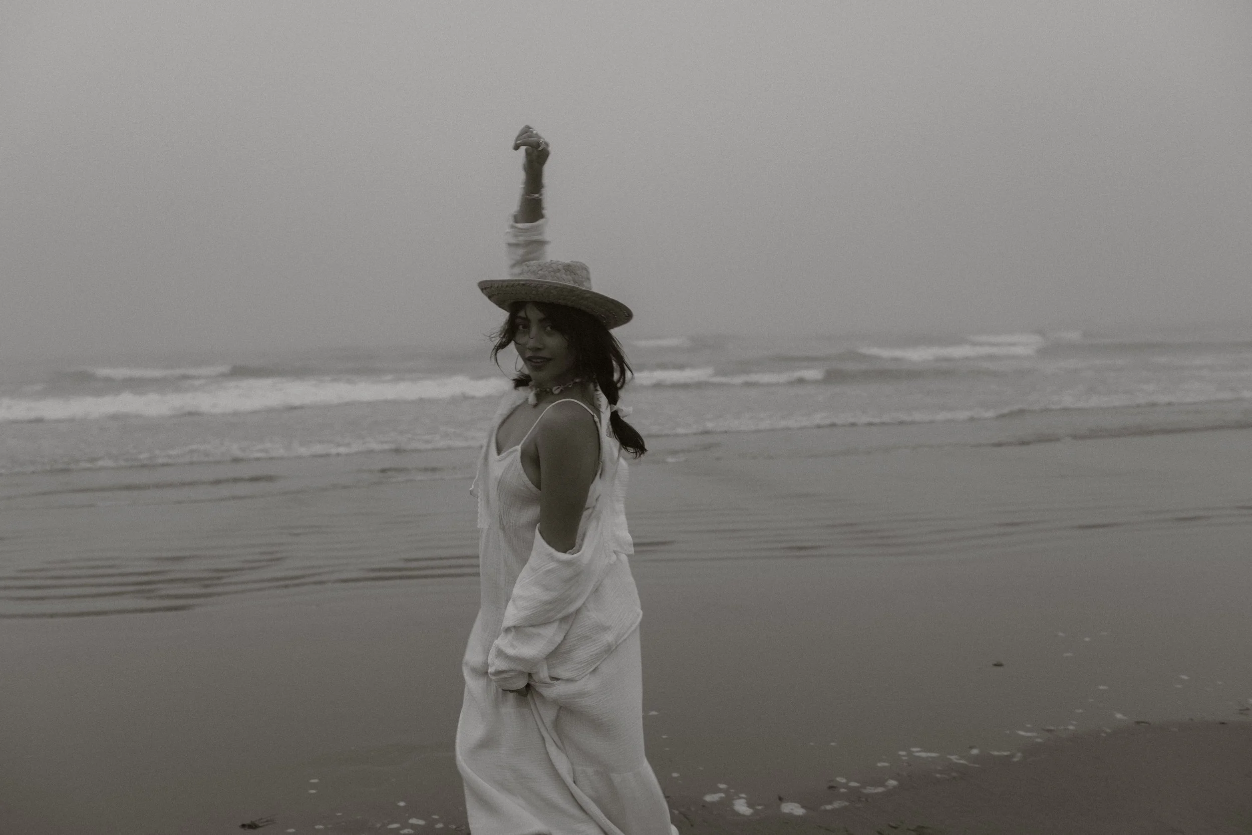 A woman wearing a hat and white dress standing on the beach near the ocean in a black-and-white photo.