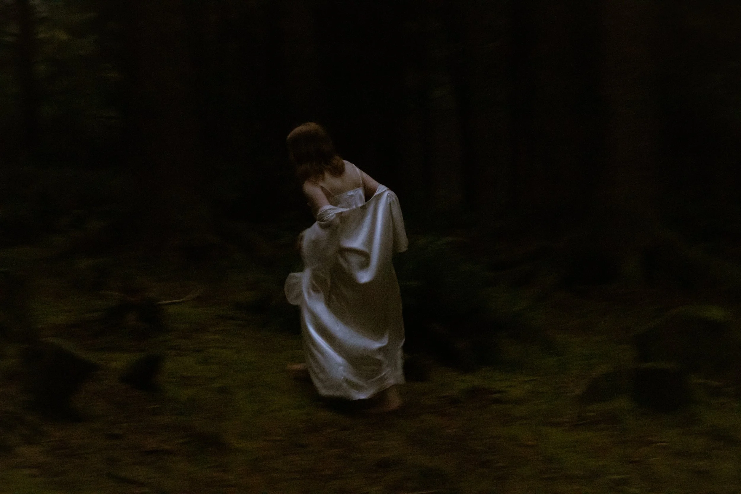 A woman in a white dress walking through a dark wooded area at night.