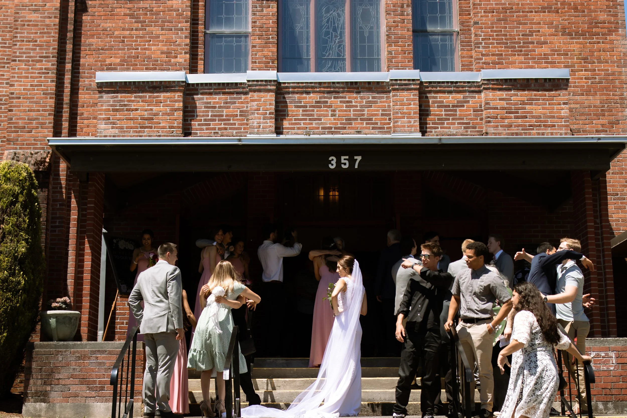 People gathered outside a brick church building for a wedding, with a bride in a white dress and veil holding a bouquet, surrounded by friends and family.