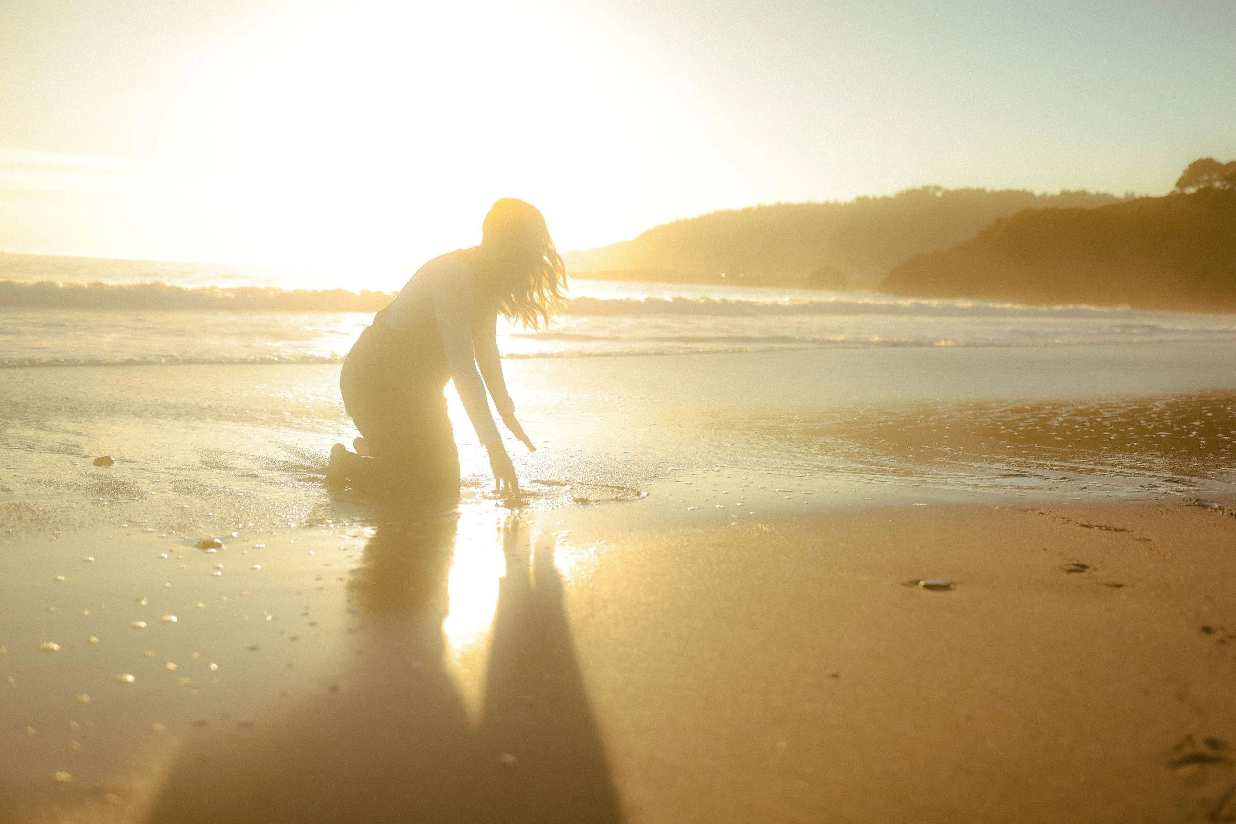 A woman kneeling on the beach at sunset, with her hands on the sand and her head down, near the shoreline with waves, mountains, and a bright sun in the background.