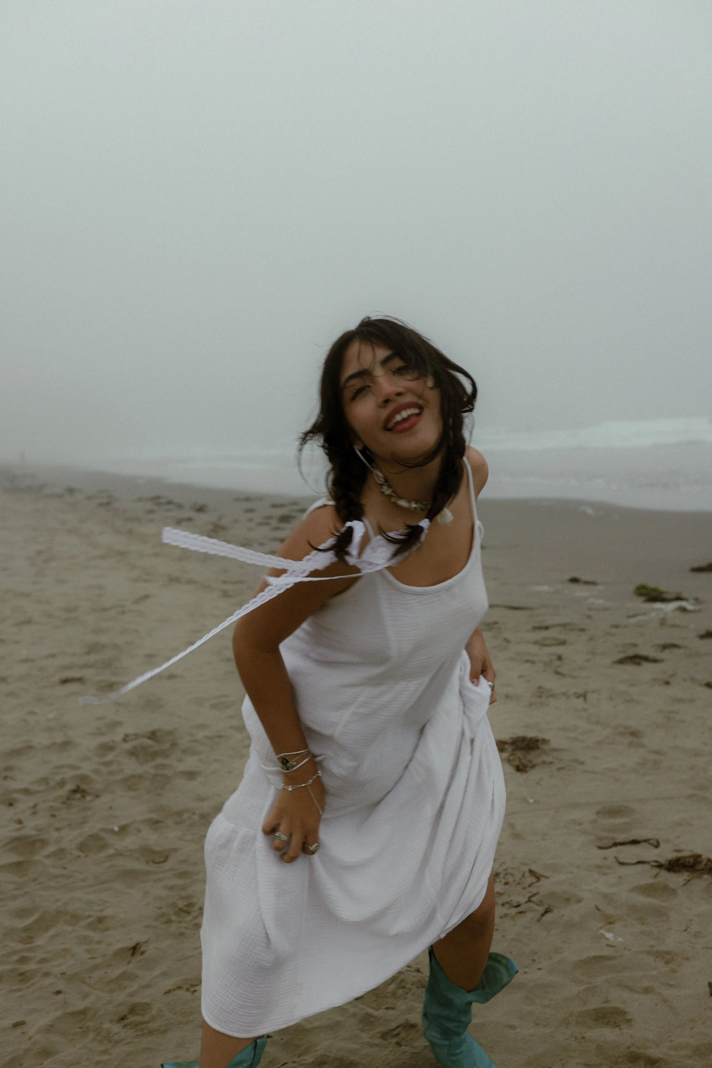 A woman in a white dress and turquoise boots smiling at the beach with sand and ocean in the background.