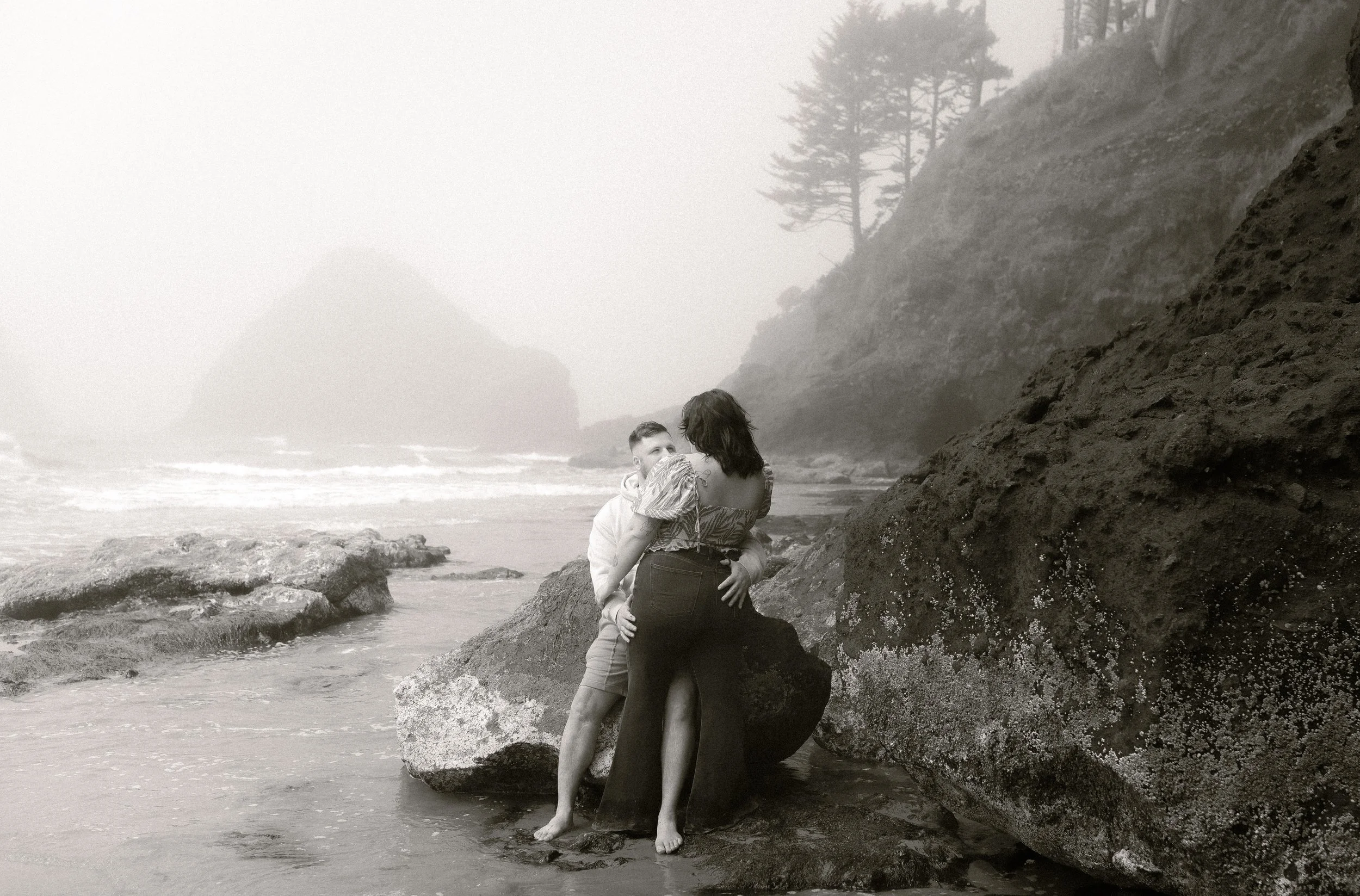 A couple embracing on a rocky beach with foggy cliffs and trees in the background.