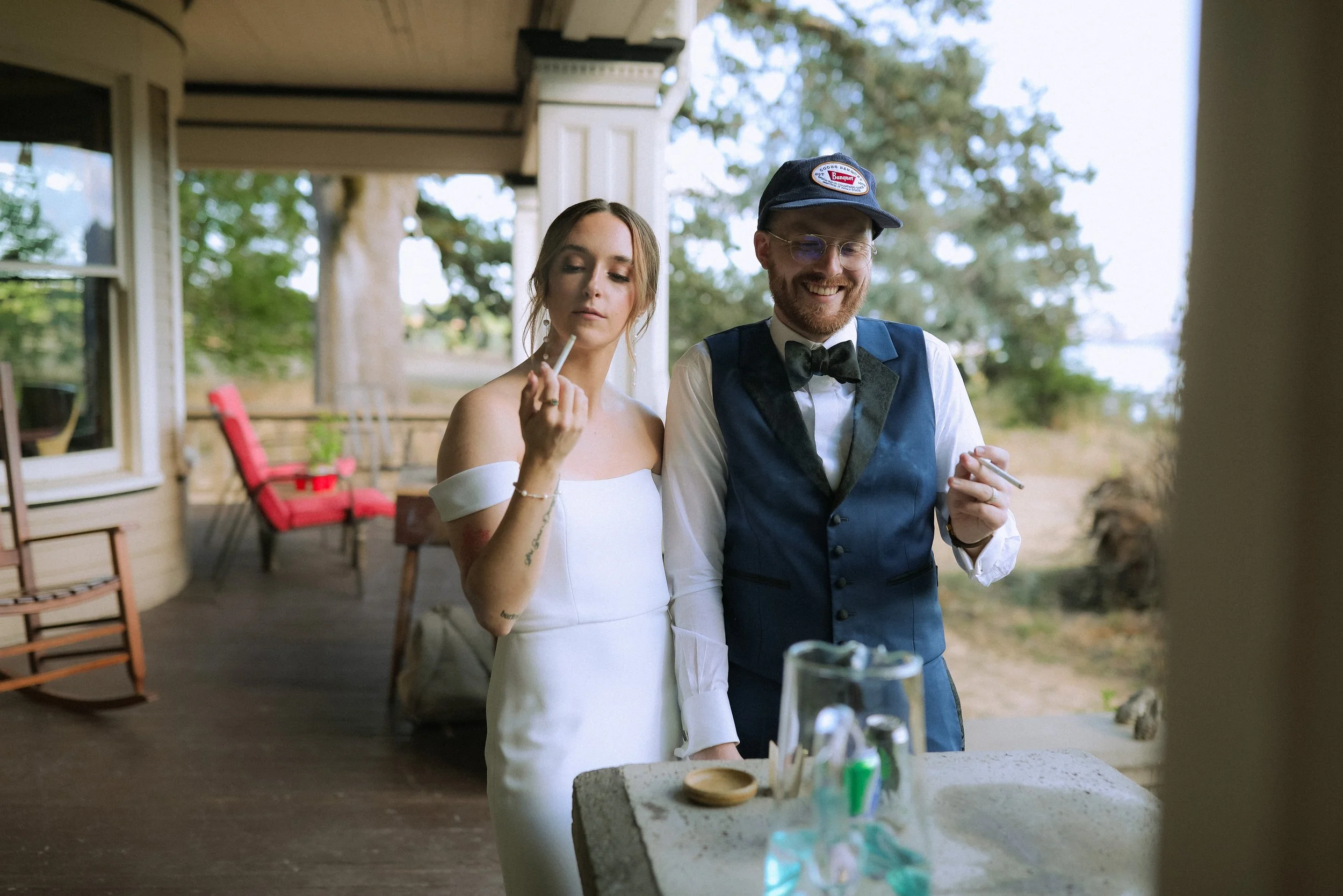 A bride and a man dressed as a butler or waiter standing outdoors on a porch, both holding cigarettes. The bride is in a white off-shoulder wedding dress with her eyes closed, and the man is in a tuxedo vest, white shirt, bow tie, and a cap, smiling.