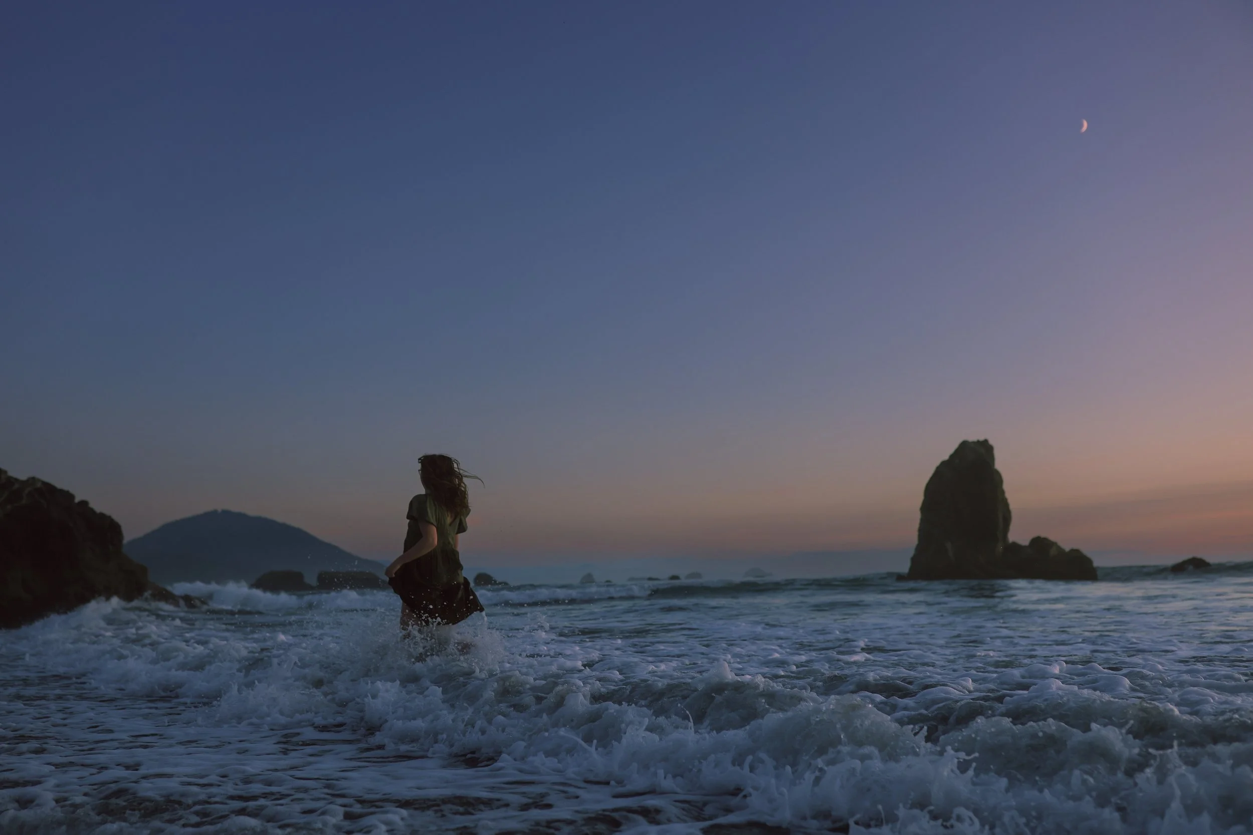 A woman walking in the ocean waves at sunset with a mountain and large rock formations in the background and a crescent moon in the sky.