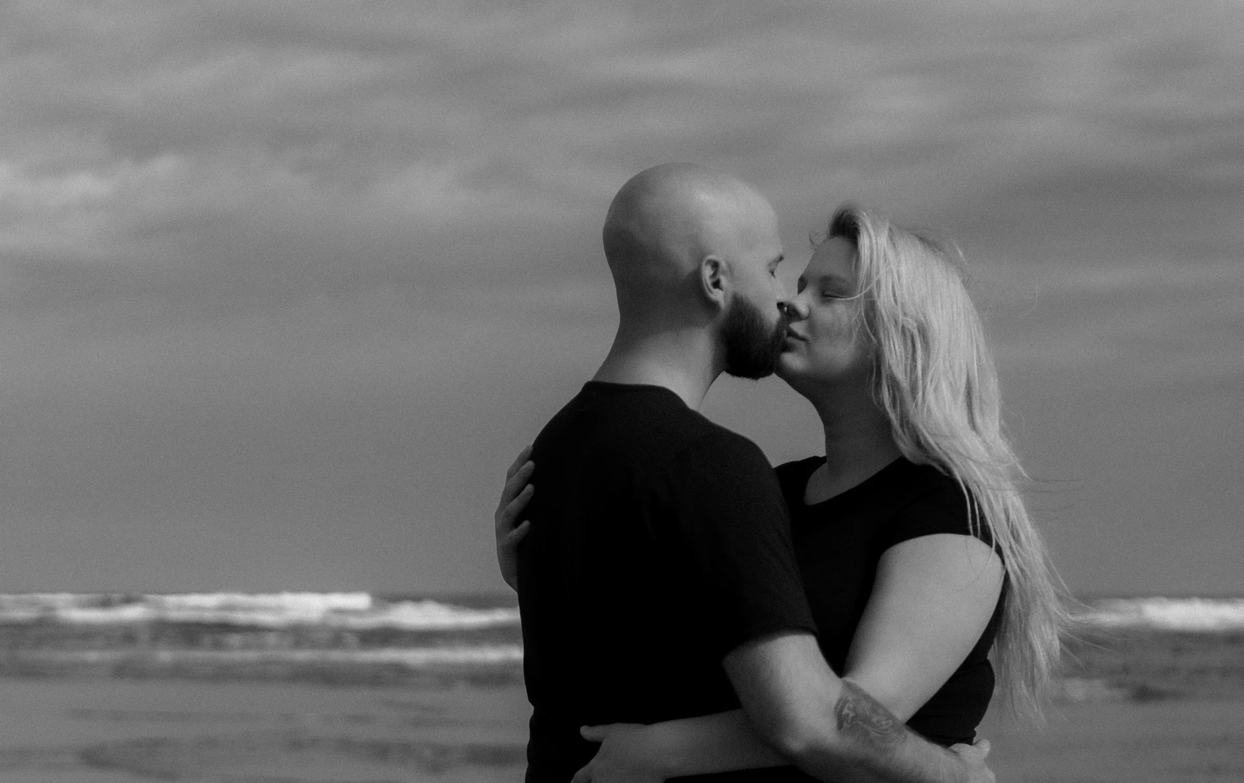 A black and white photo of a couple on a beach kissing, with the ocean and cloudy sky in the background.