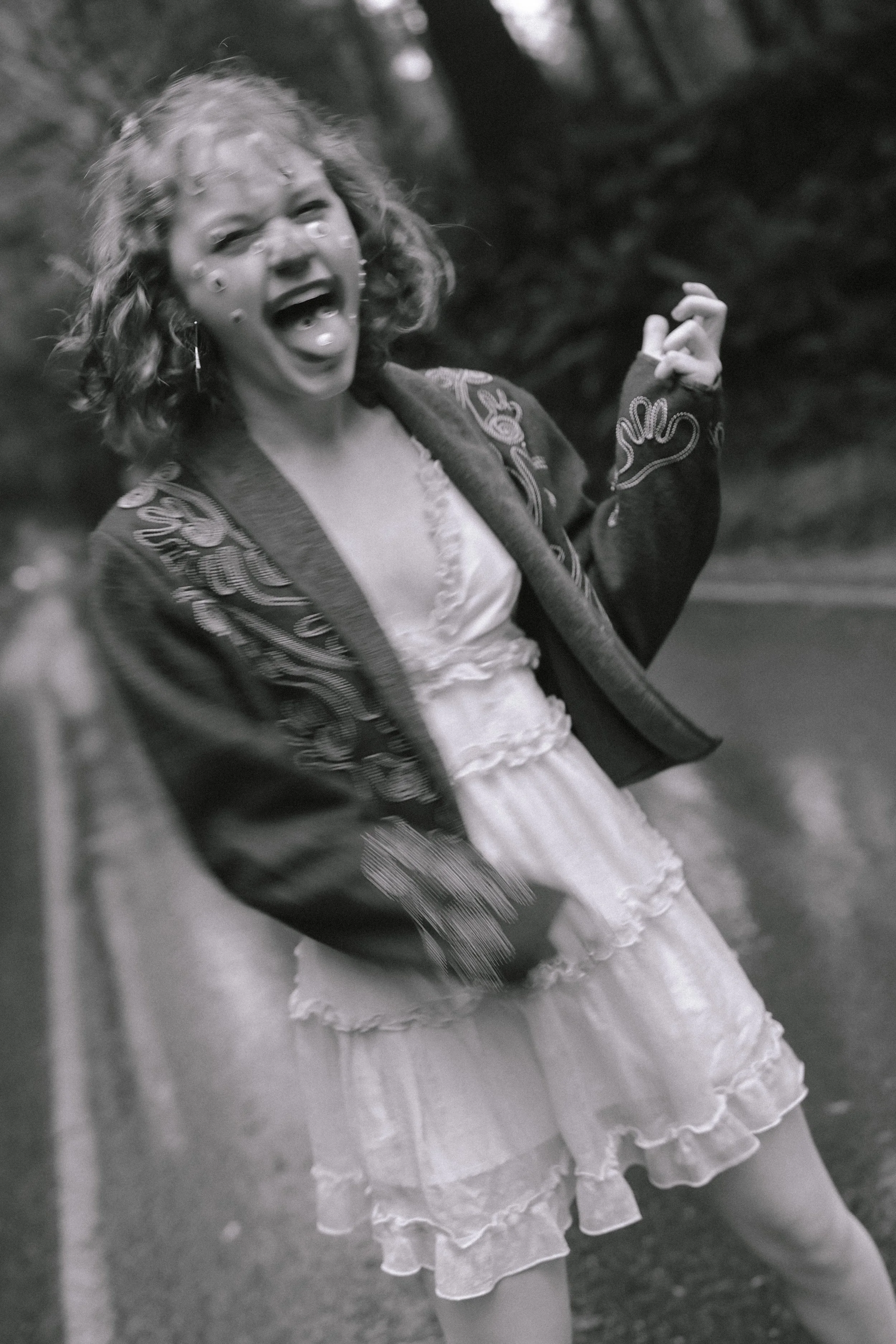 A young girl with curly hair, wearing a dark embroidered jacket and a light ruffled dress, standing outdoors near a road, laughing with her mouth open and eyes closed.