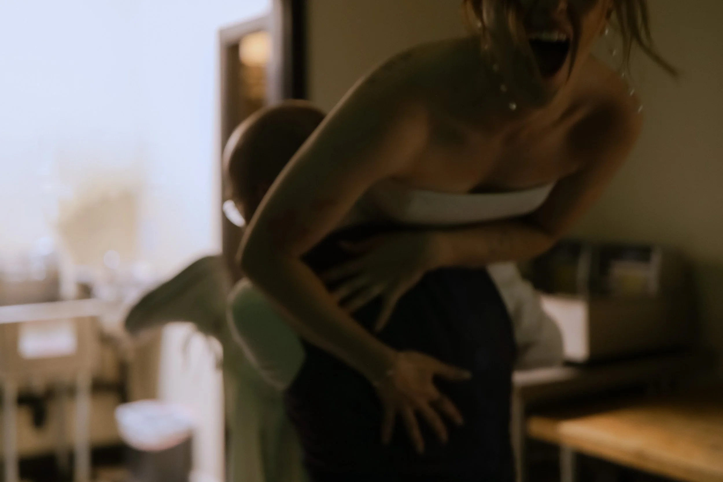 A woman laughing and holding her stomach, standing in a dimly lit room with another person behind her, possibly a child.