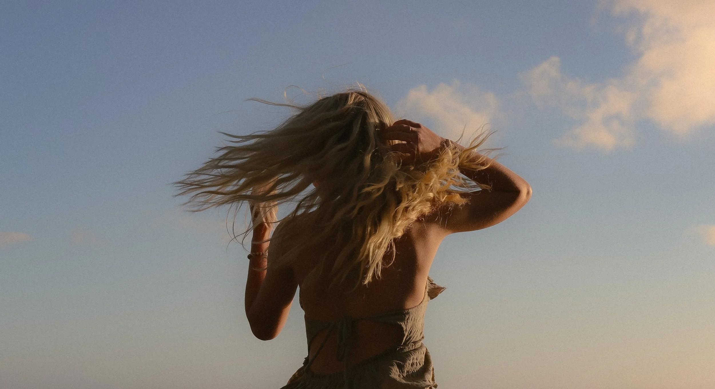 Woman with long blonde hair outdoors against a blue sky with clouds, touching her hair.