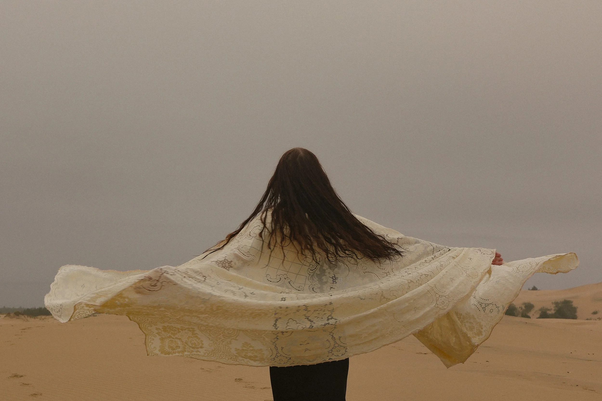 A woman with long hair standing in a desert, holding a large, flowing, patterned cloth or scarf spread out like wings.