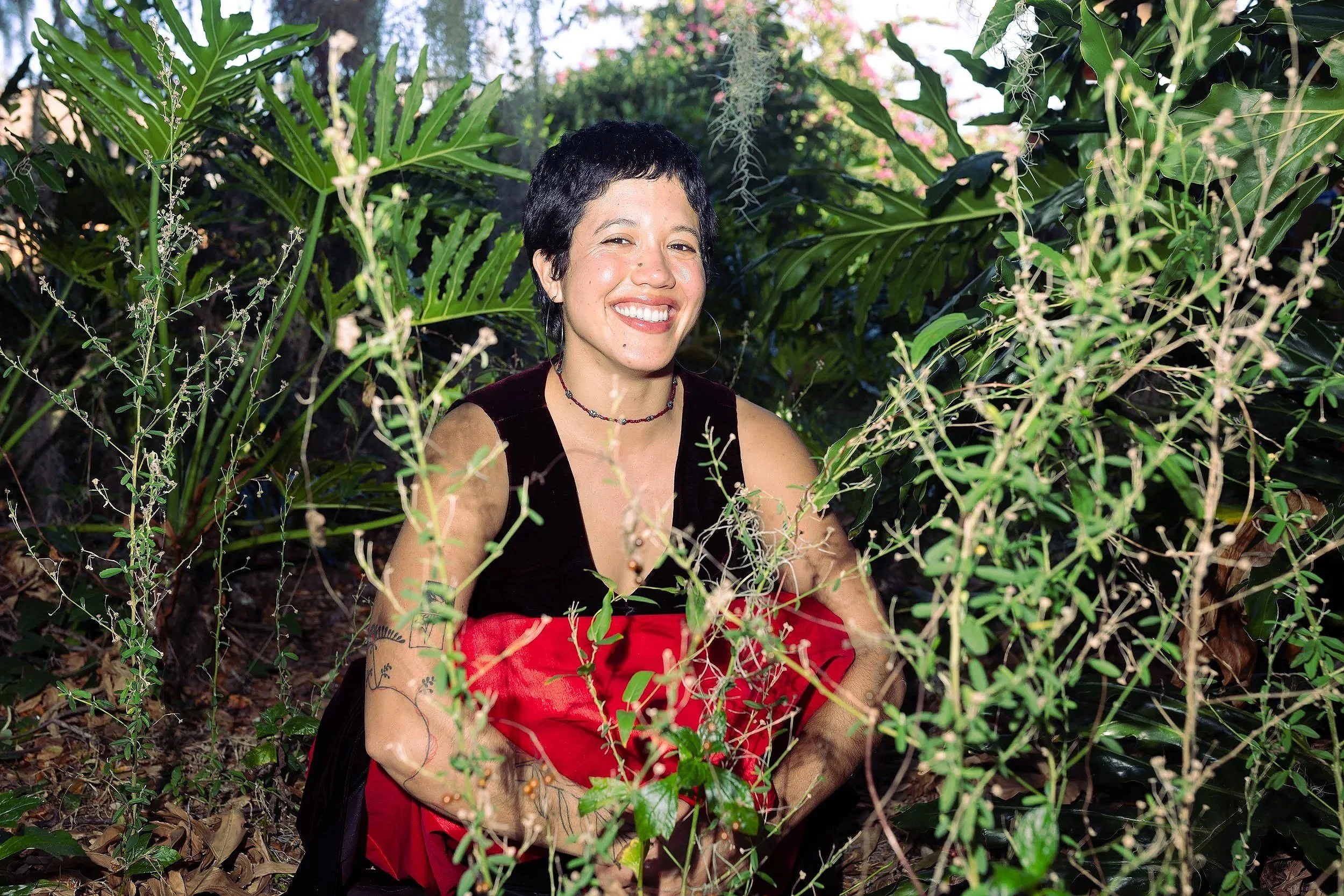 A woman with short black hair, smiling, sitting in a lush green garden surrounded by plants and foliage.