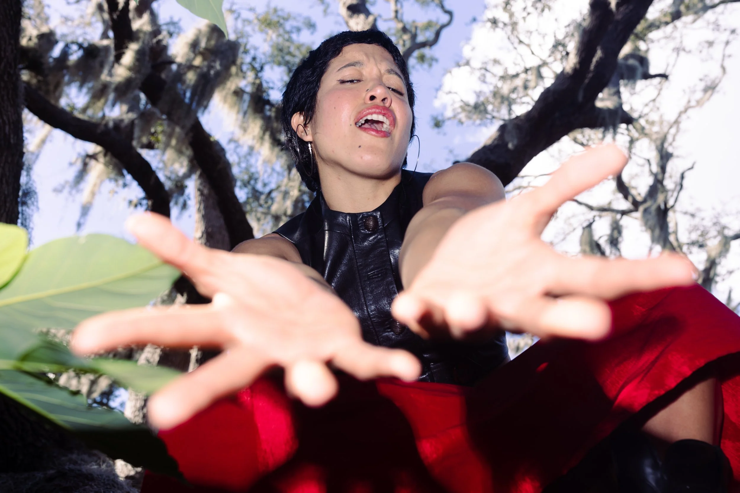 A woman with short black hair reaching towards the camera with both hands, wearing a black leather top and a red skirt, outdoors with trees and blue sky in the background.