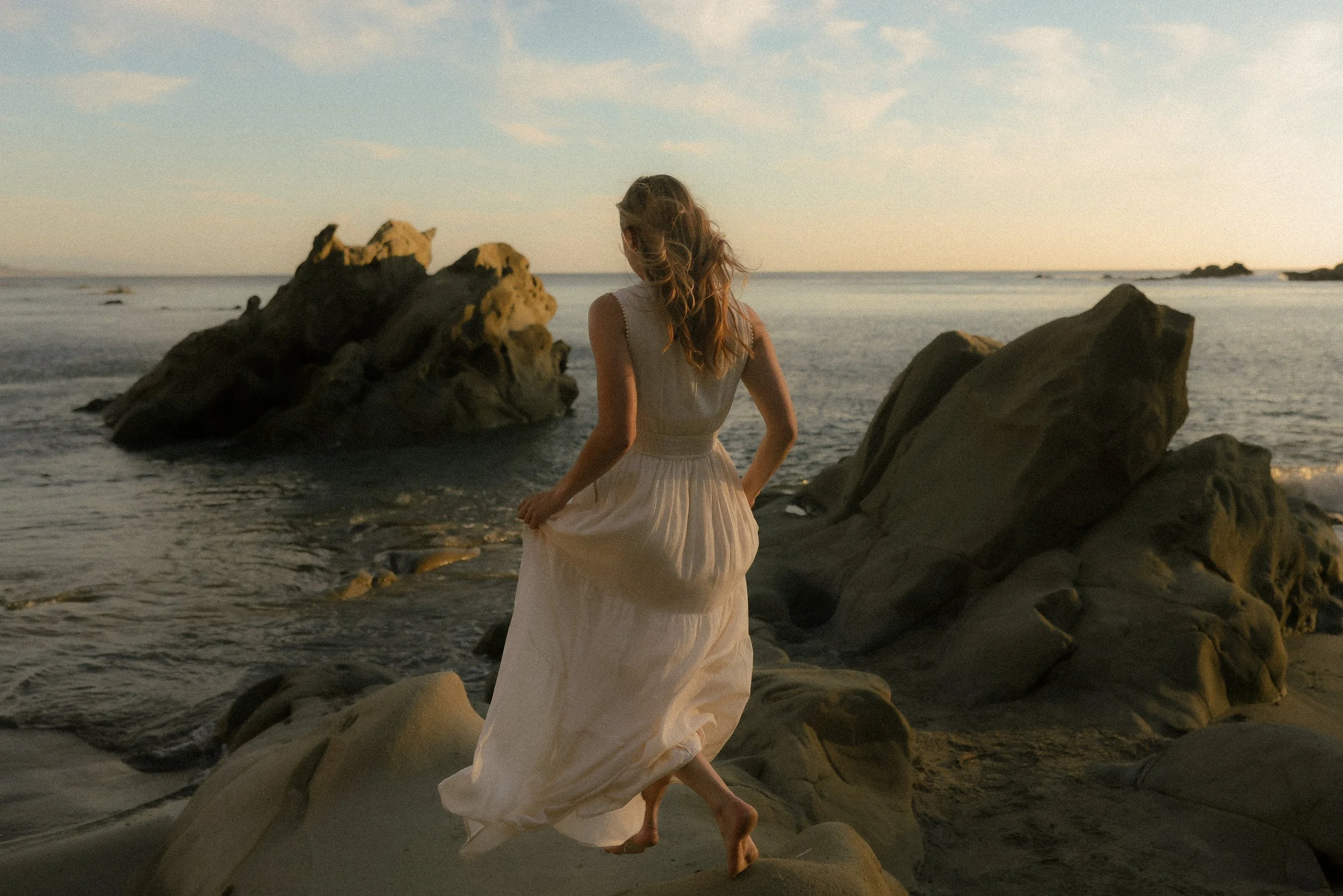 A woman in a white dress walking on rocks at the beach during sunset.