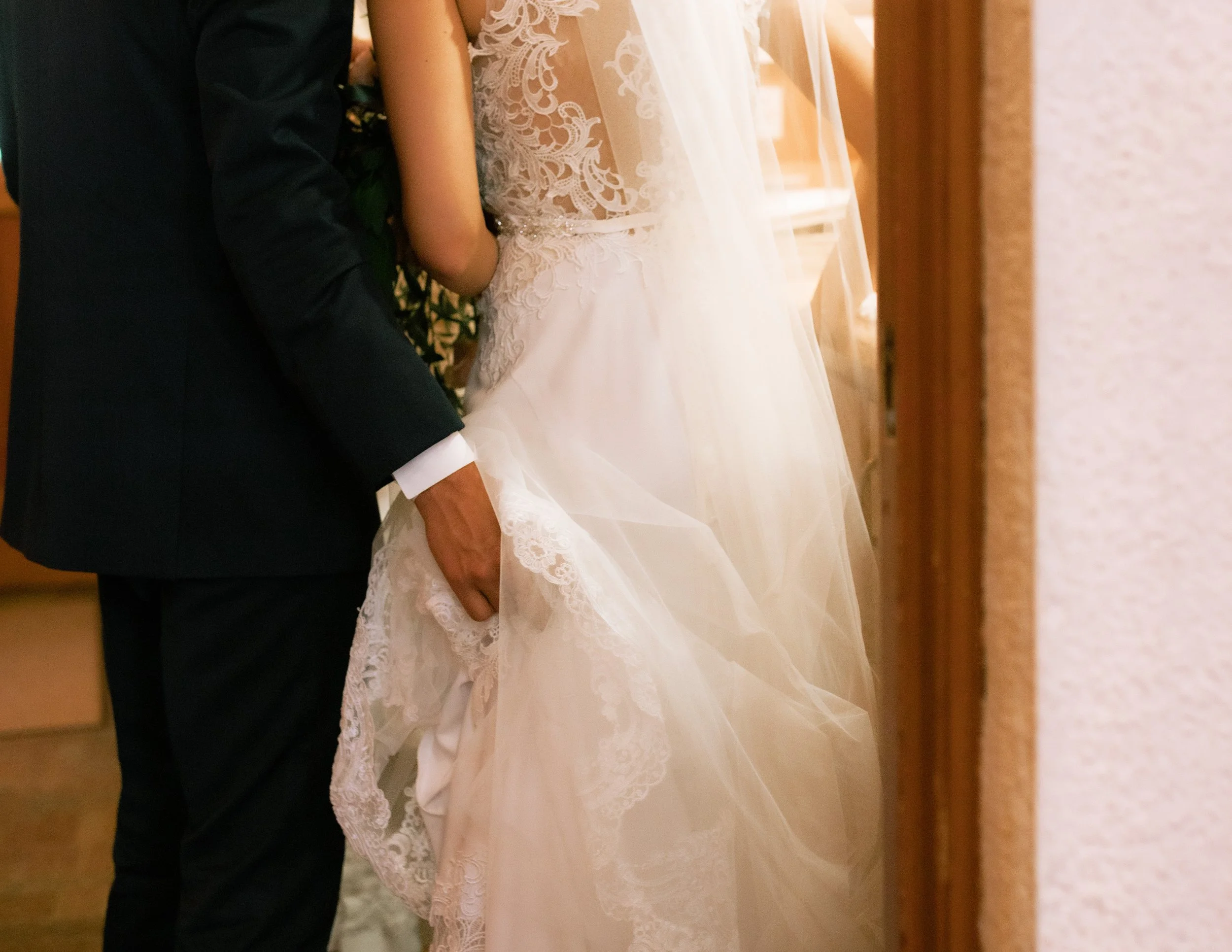 A bride in a white lace wedding gown and a groom in a black suit sharing a moment, with the groom holding the bride's dress, in an indoor setting