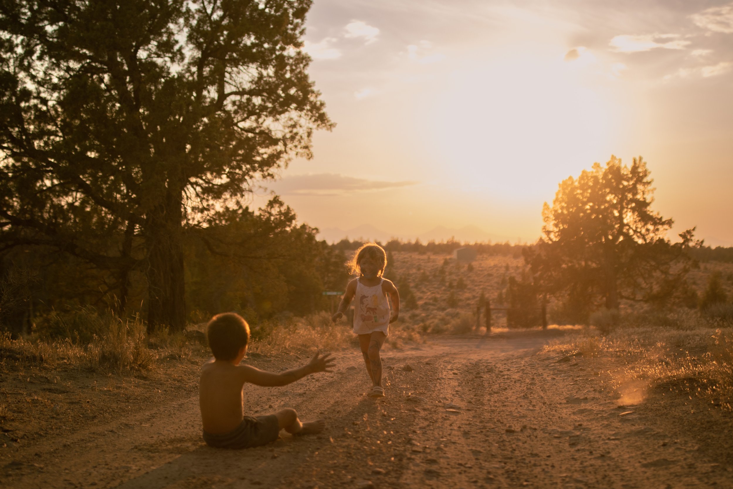 Two children playing on a dirt trail in a rural area during sunset, with trees and a fence in the background.