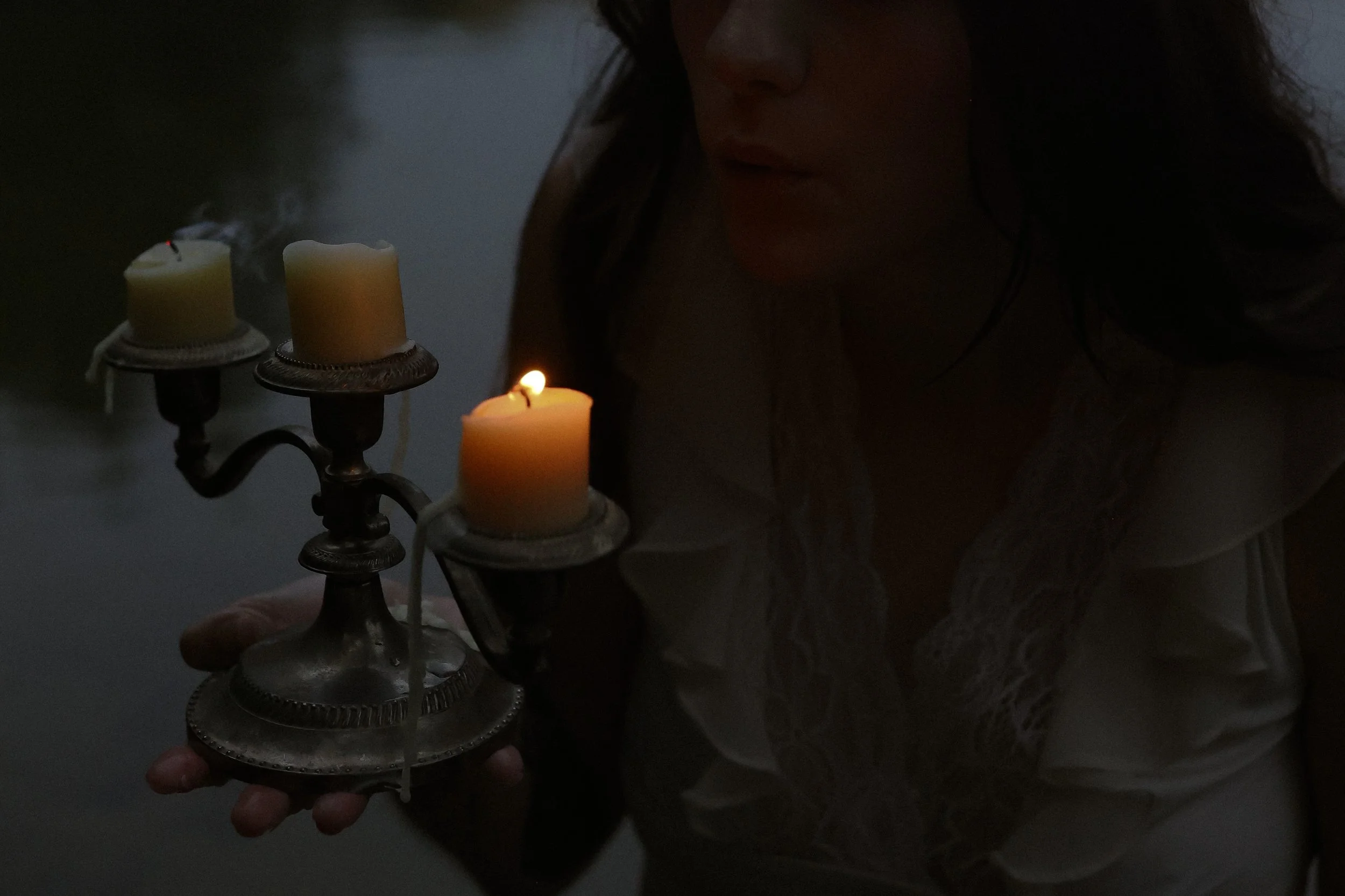 A woman holding a vintage metal candle holder with three lit candles, in a dark environment.