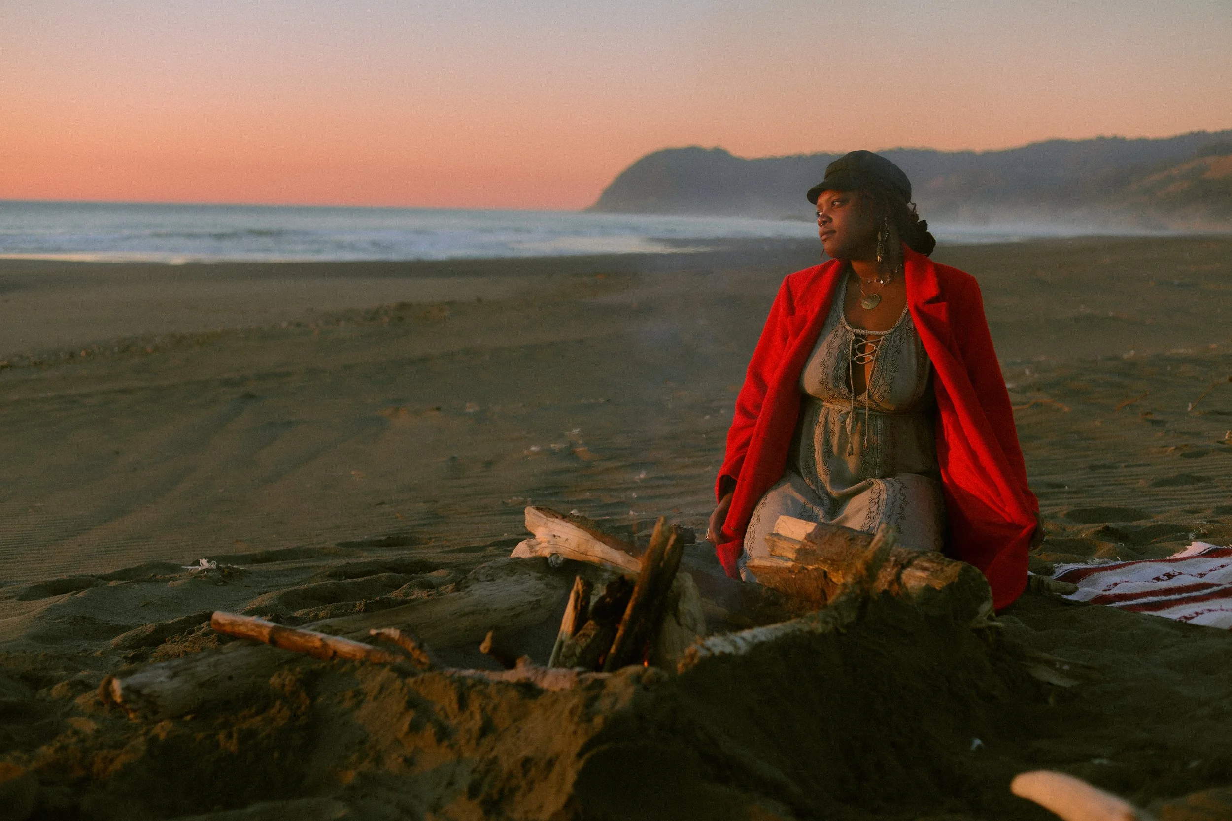 A woman in a red coat and gray dress sits on the beach near a campfire during sunset, with hills and the ocean in the background.